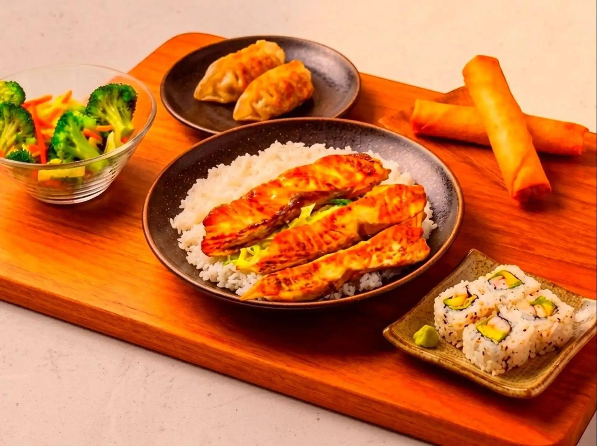 Assorted Asian dishes including grilled chicken over rice, sushi rolls, salad with broccoli and carrots, gyoza dumplings, spring rolls, and a dipping sauce on a wooden serving board.