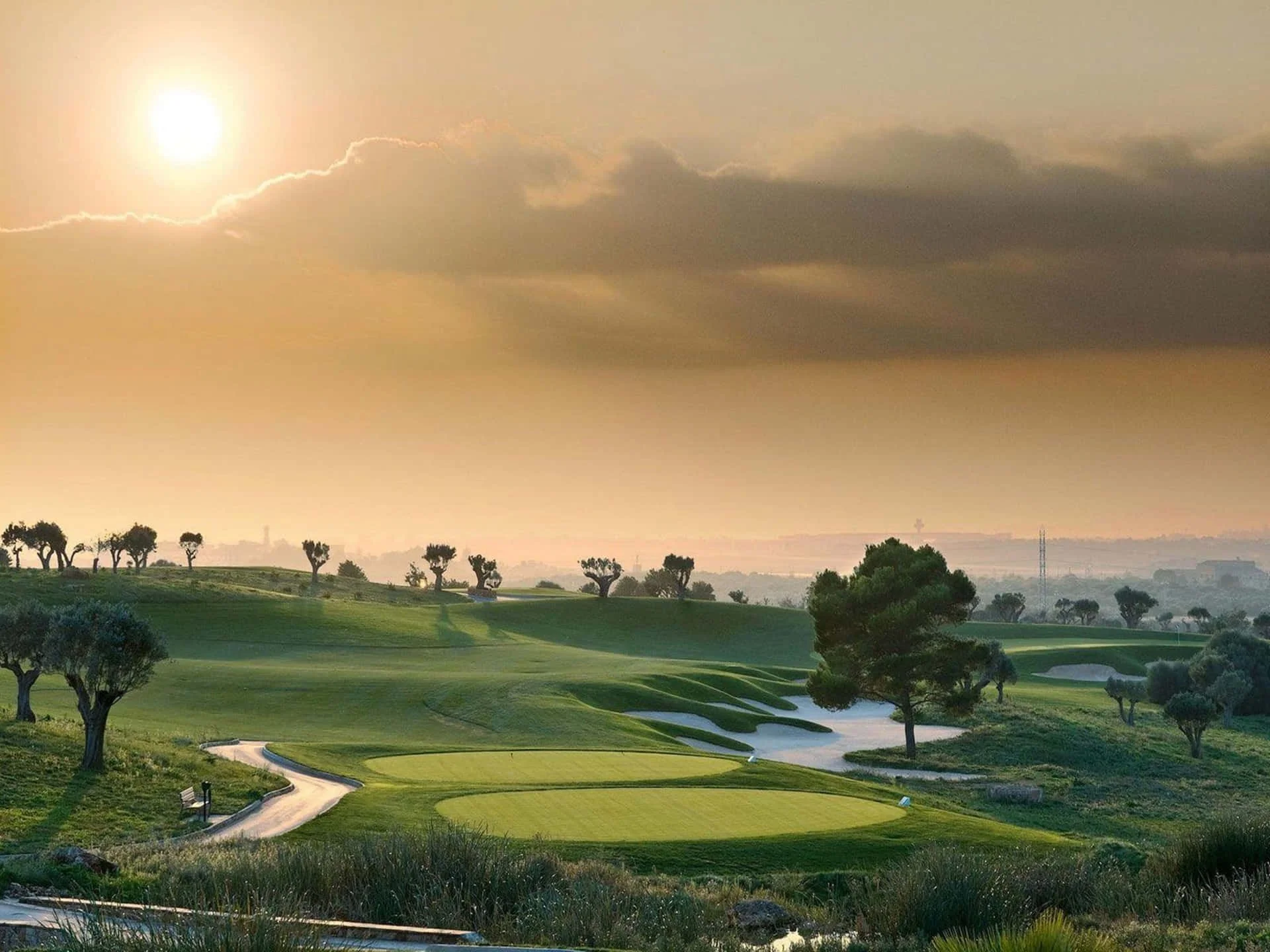 A golf course landscape at sunset with trees, neatly trimmed grass, a sand trap, and a winding path.