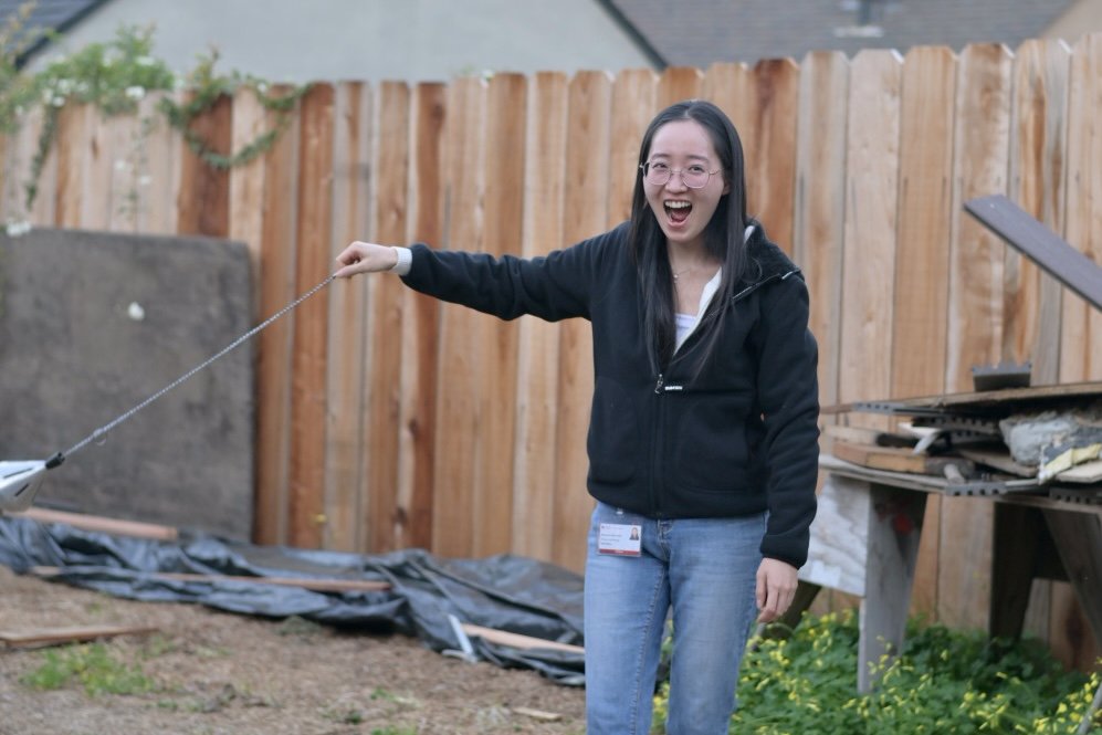 A woman with long black hair, glasses, and a black jacket learning to swing a church incense burner (a thurible), smiling and laughing outdoors in a backyard.