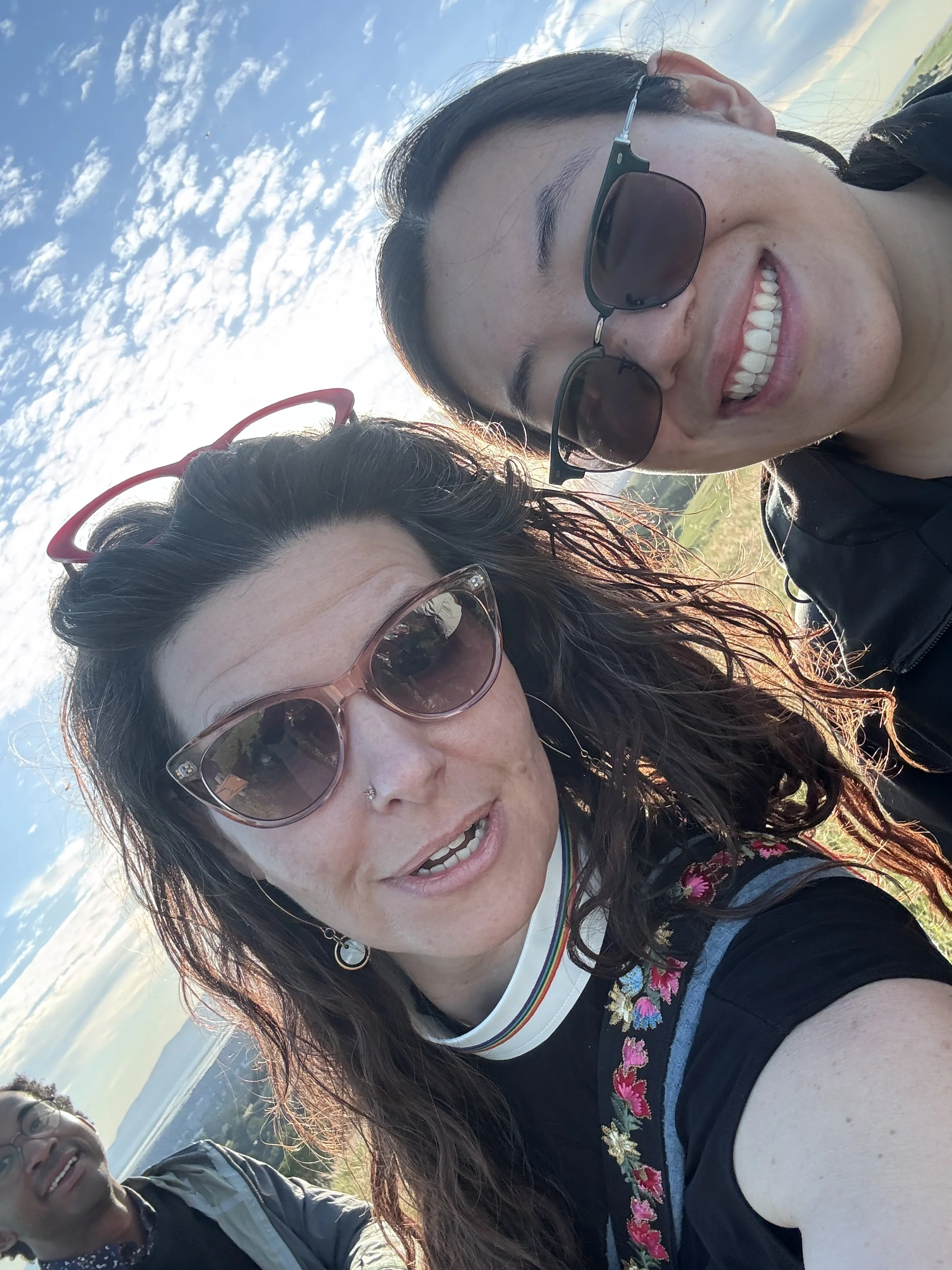 A woman with long dark hair, wearing sunglasses and earrings, smiling while taking a selfie with another woman in the background during sunrise, with a landscape and a sky filled with scattered clouds.