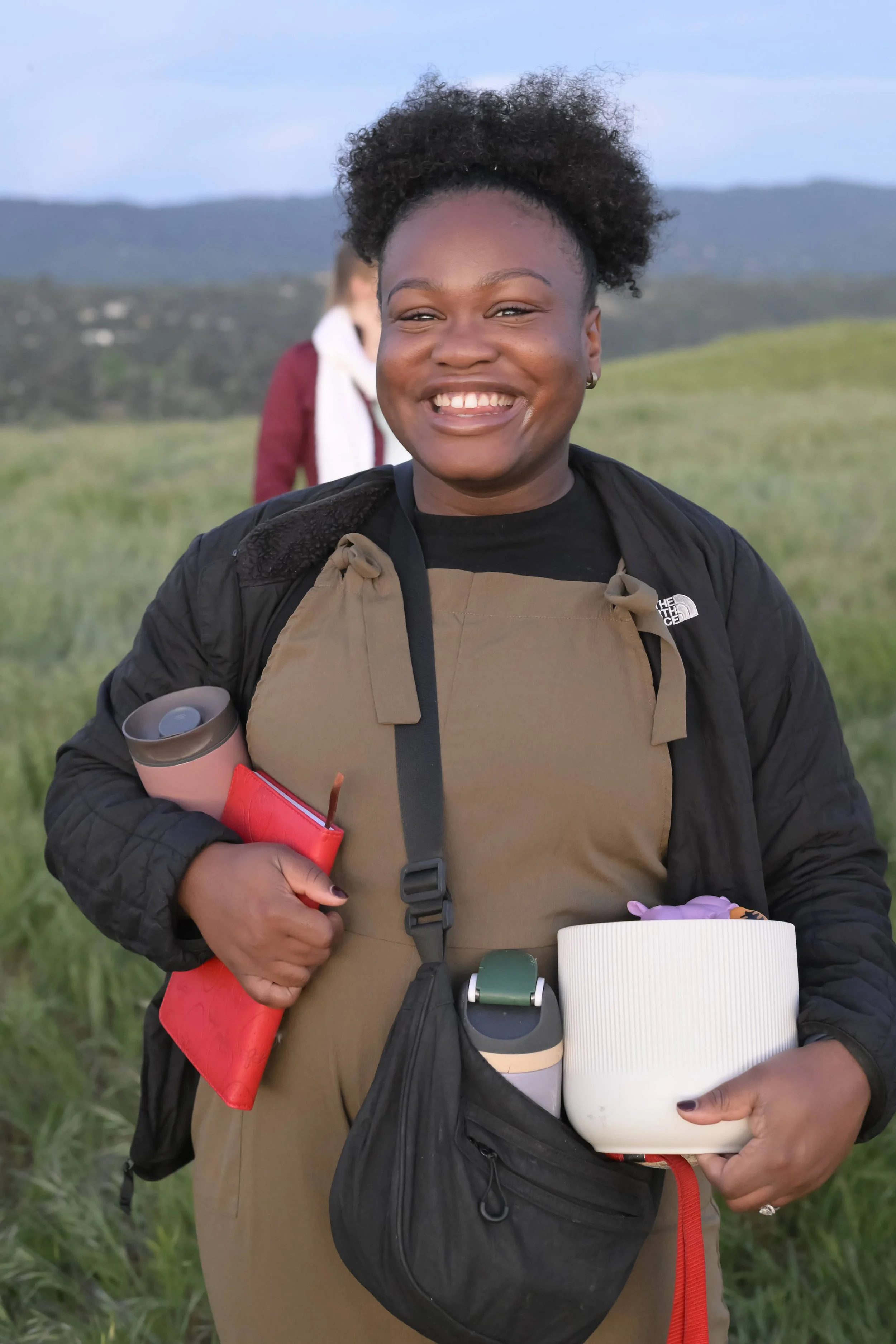 Close-up of a smiling woman with dark curly hair, holding a reusable water bottle and a white cylindrical container, wearing a black shirt and tan overalls, standing outdoors in a grassy area with hills in the background.