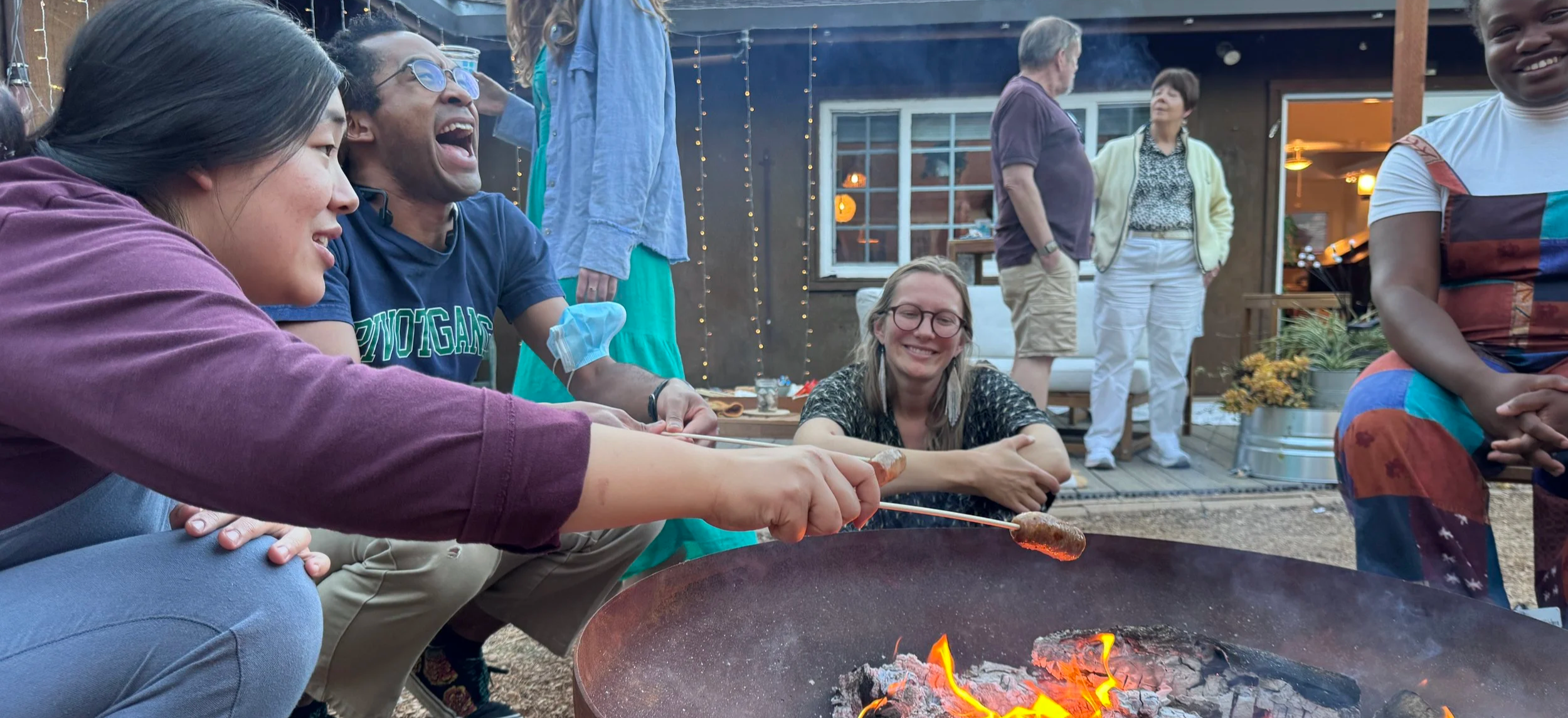 Group of friends roast marshmallows and sausage over a fire pit at an outdoor gathering during the evening. They are smiling and enjoying each other's company.