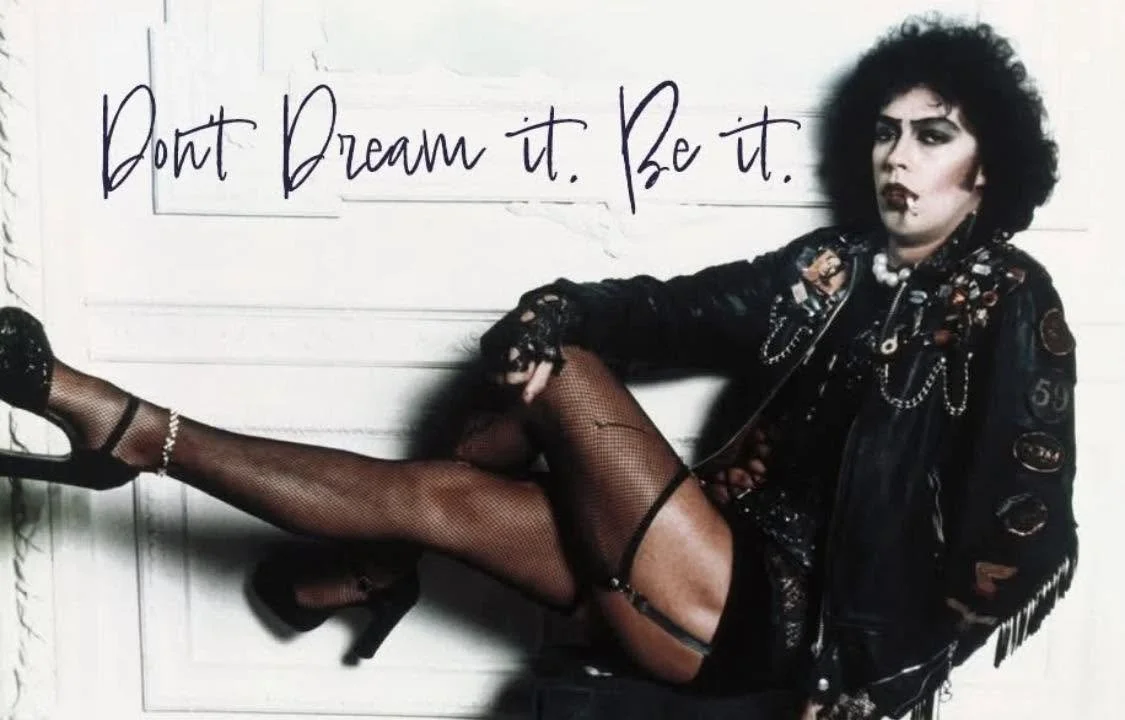 Actor Tim Curry, dressed in platform heels, fishnet stockings, hot shorts, leather fringe jacket and pearls, heavy drag makeup, with cigarette in his mouth. The words "Dont Dream it. Be it" written in the white space above his pose.