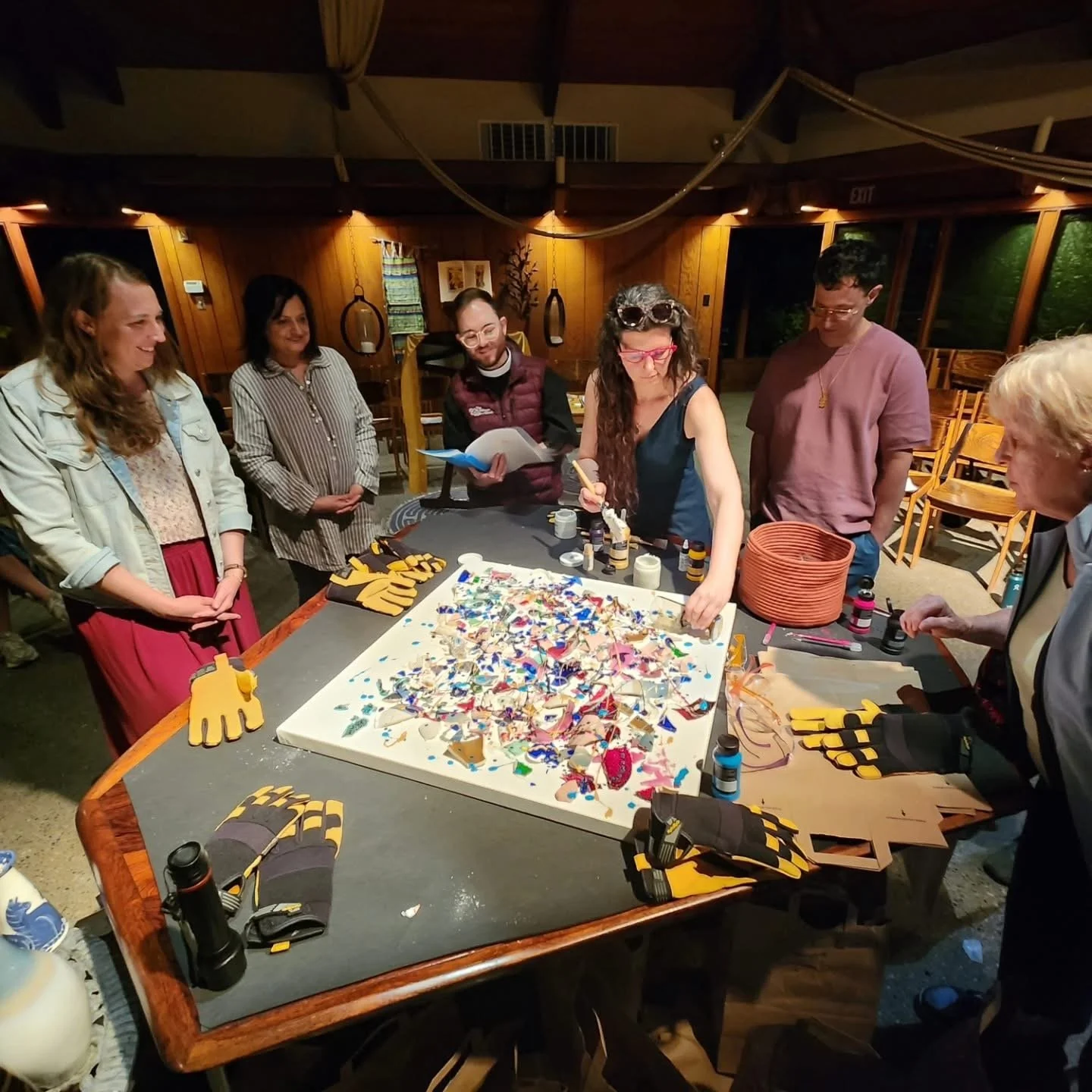 Group of people gathered around a table with a mosaic art project, tools, and supplies, engaging in a creative activity in a warmly lit indoor environment.