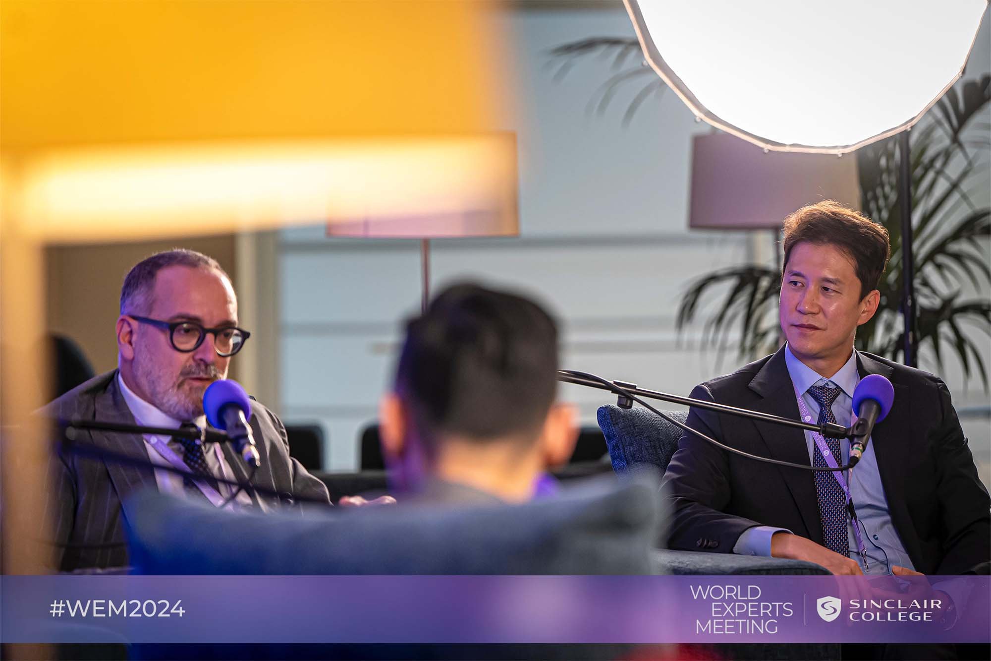 Two men wearing business suits and glasses are seated at a table with blue microphones in front of them, engaged in a discussion during the World Experts Meeting at Sinclair College. There are studio lights and large plants in the background.