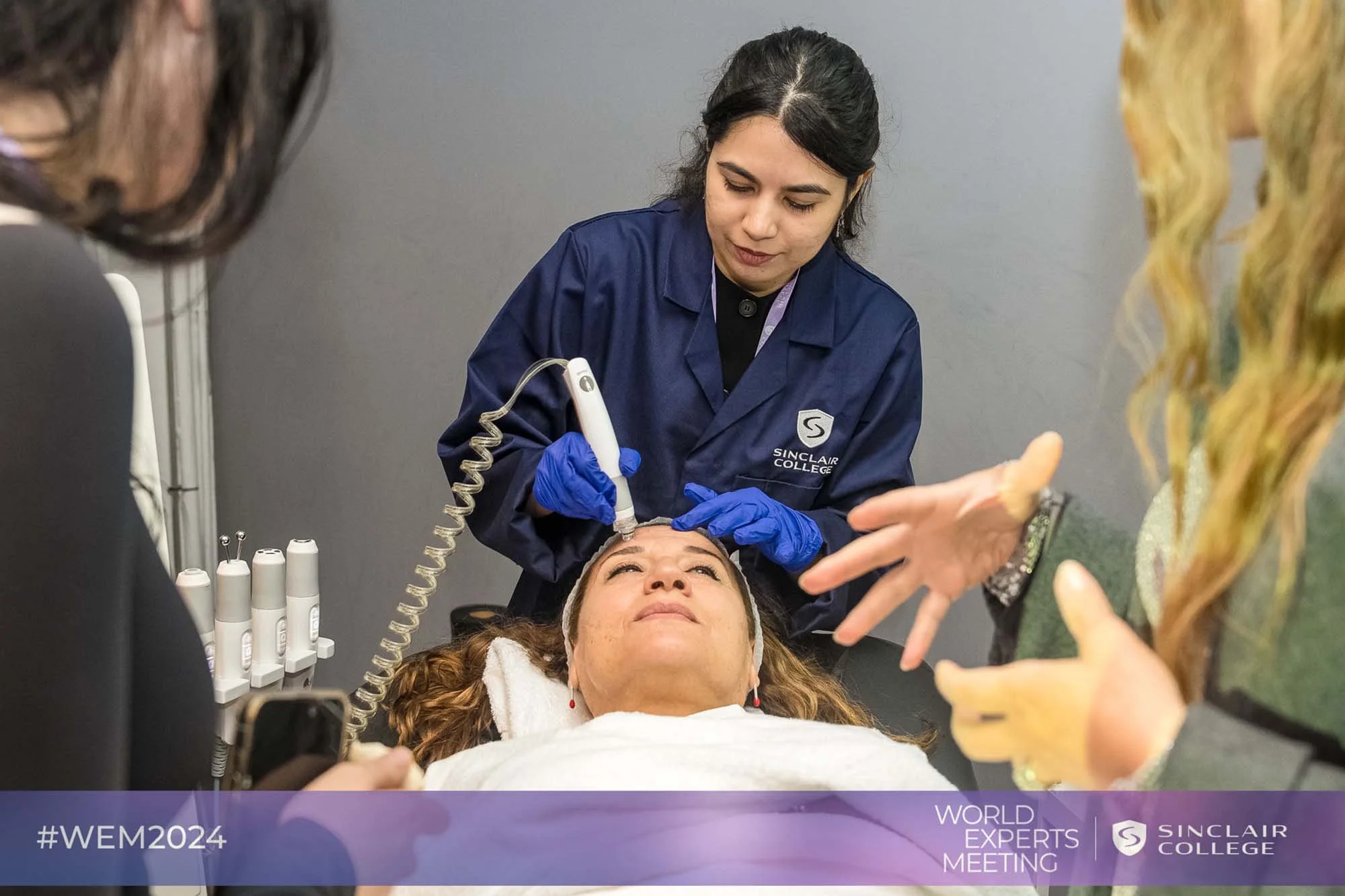 A woman with curly hair receiving a cosmetic treatment from a healthcare professional in a clinic, with other people engaged in conversation nearby. The scene is part of a World Experts Meeting at Sinclair College, as indicated by the purple banner at the bottom of the image.