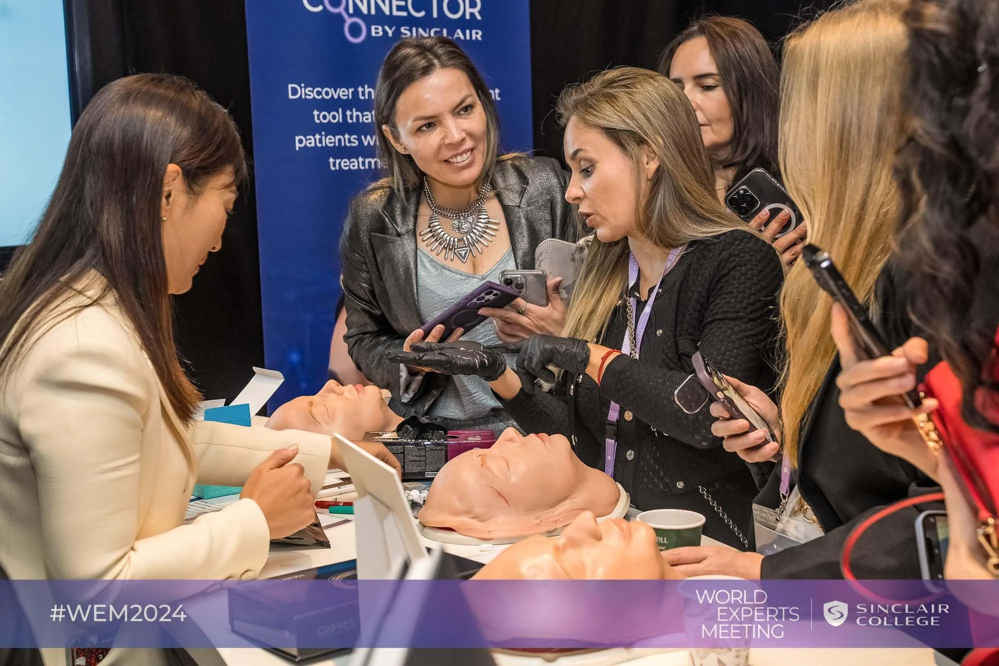 A group of women gathered around a table at a medical demonstration, watching a woman perform a demonstration on a prosthetic head. They are at a conference, with a banner in the background indicating it is a World Expert Meeting at Sinclair College.