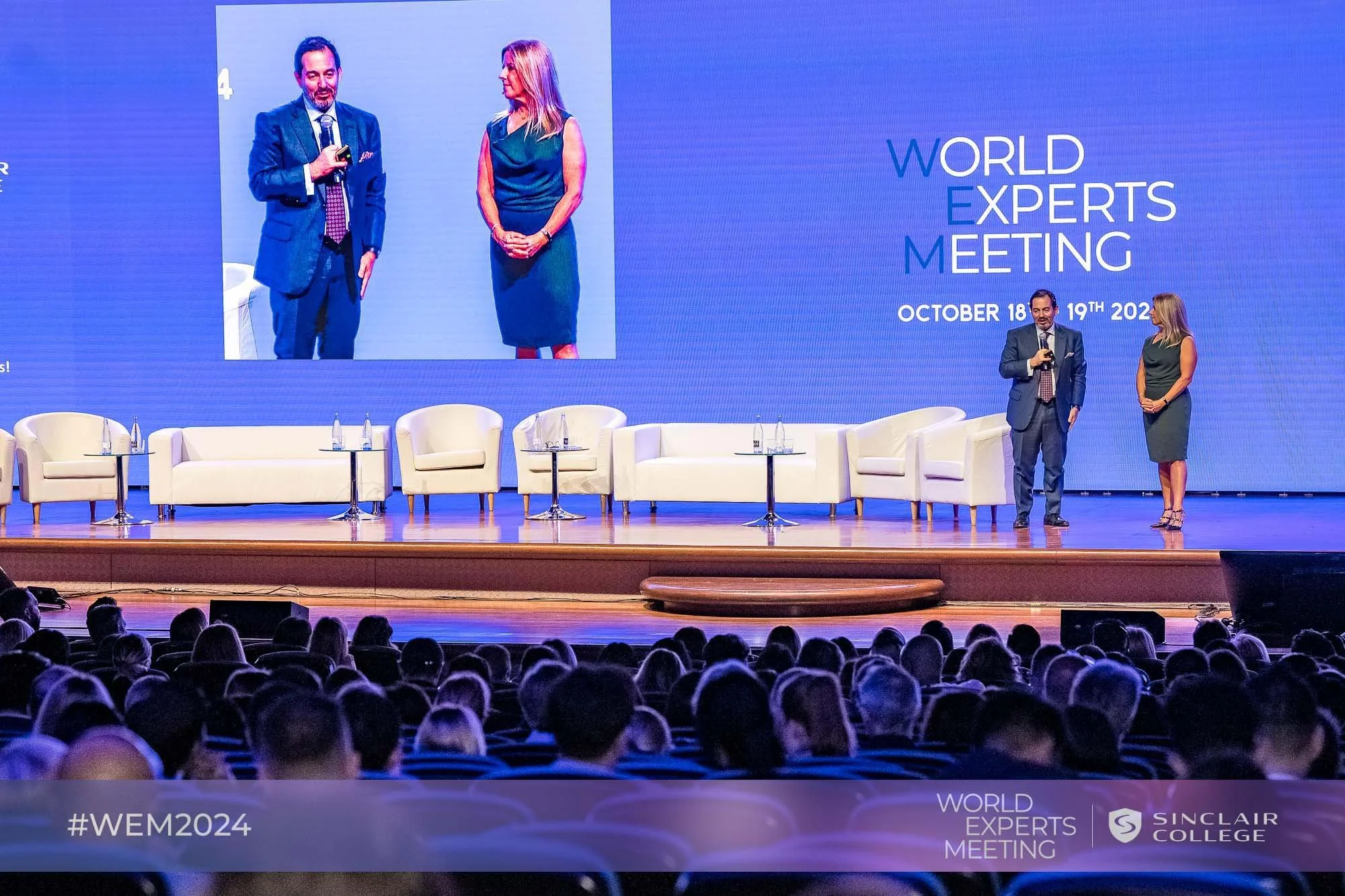 Two speakers on stage at a conference, with one man holding a microphone and a woman standing beside him, a large screen displaying the event's name and date behind them, and an audience in front.