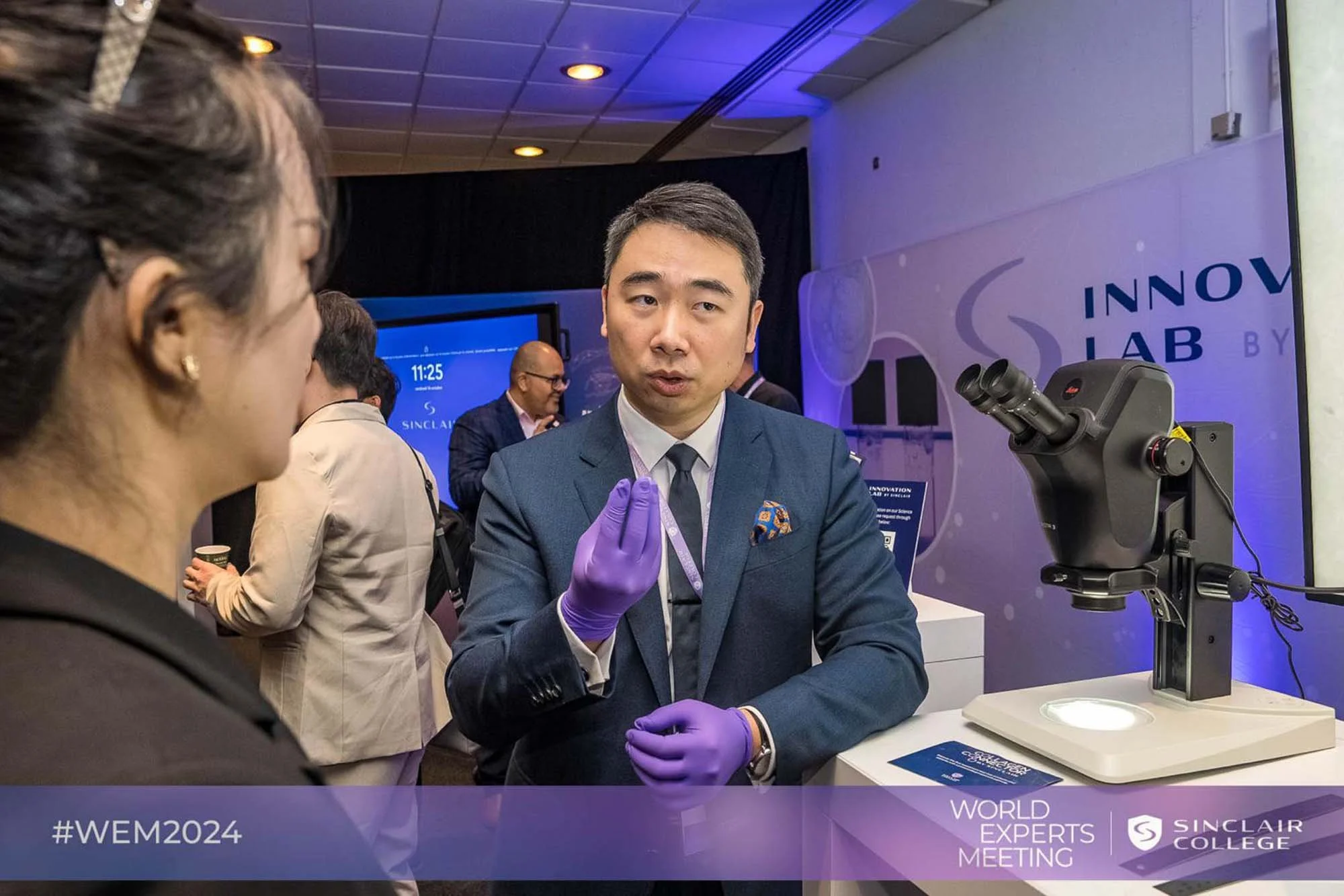 A man in a suit and purple gloves demonstrating a microscope to a woman at a science conference. The background features a sign reading 'INNOV LAB BY SINCLAIR COLLEGE' and other attendees.