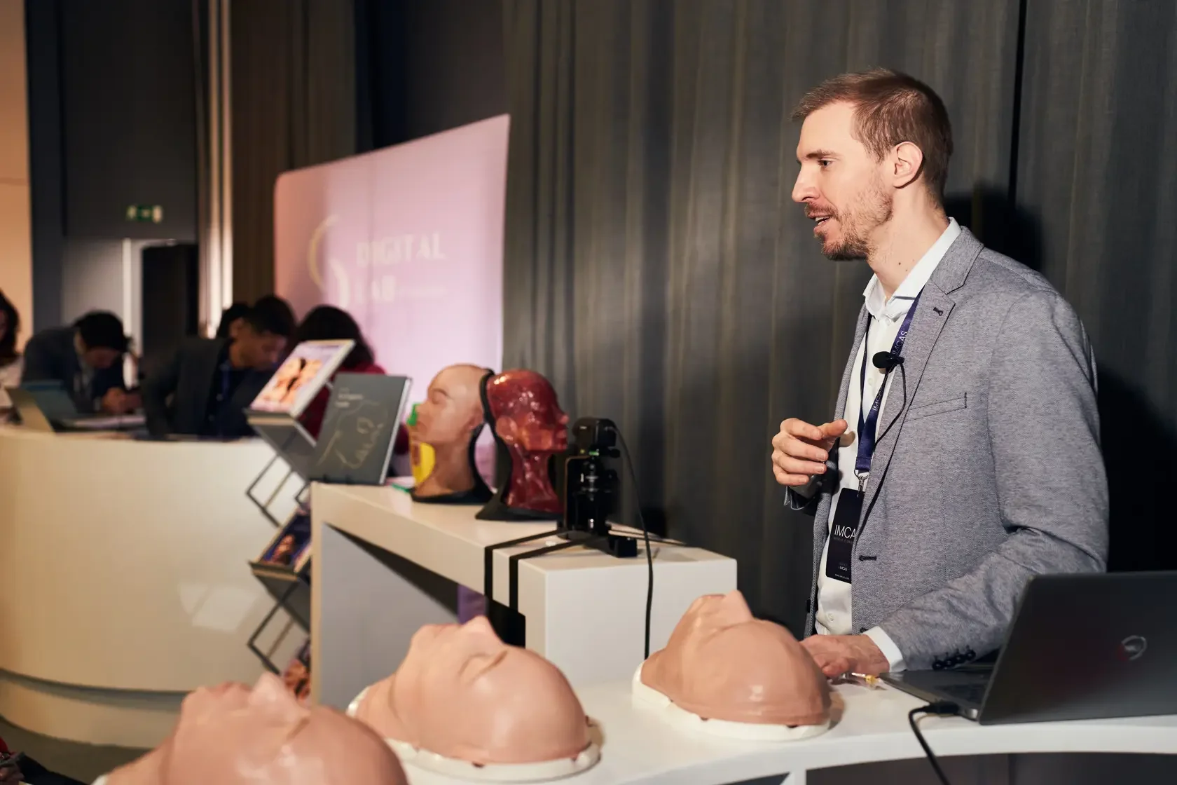 A man in a gray blazer stands at a booth with anatomical models and exhibits, at a conference or medical exhibition, with a large screen displaying 'DIGITAL' in the background.