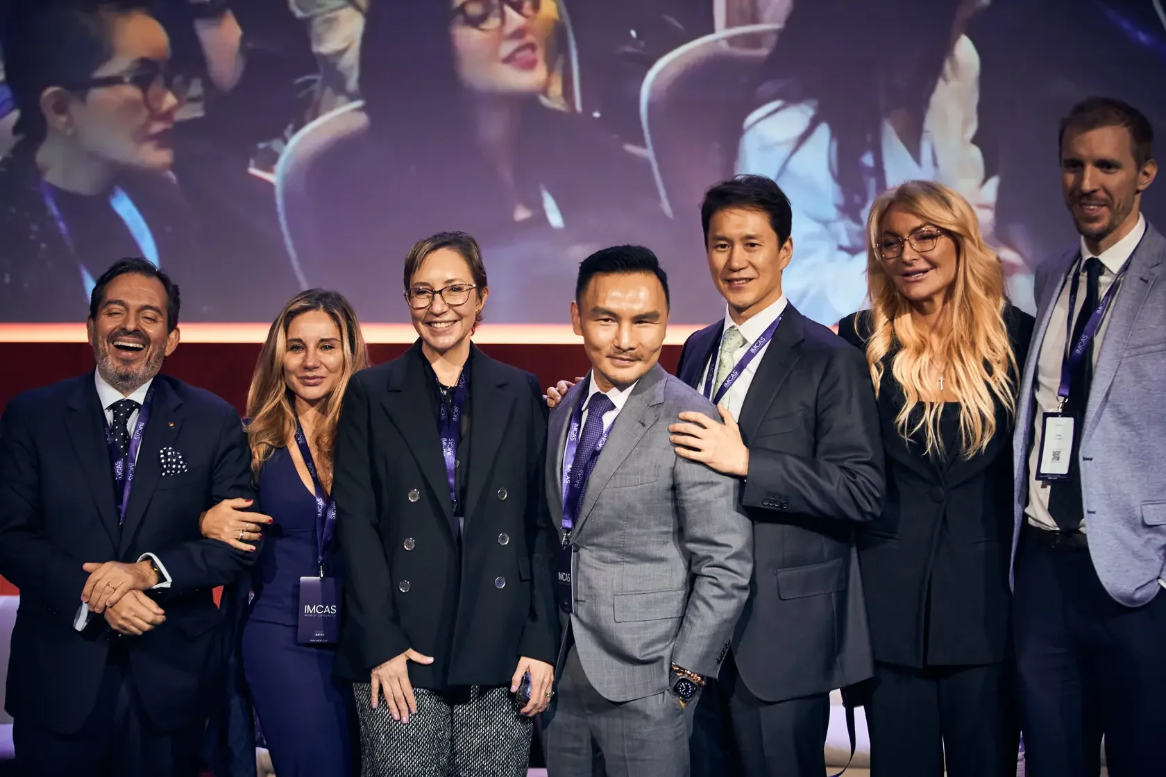 Group of eight diverse professionals dressed in formal business attire, standing on a stage with a large screen behind them displaying a similar image. They are smiling and posing for the camera at a conference or event.