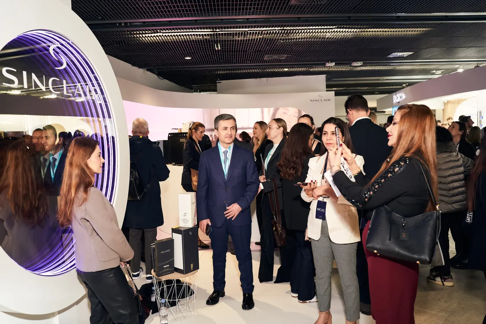 A group of business people and attendees at a conference or trade show, gathered around a booth with a sign that reads 'SINCLAIR,' engaging in conversation and taking photos.