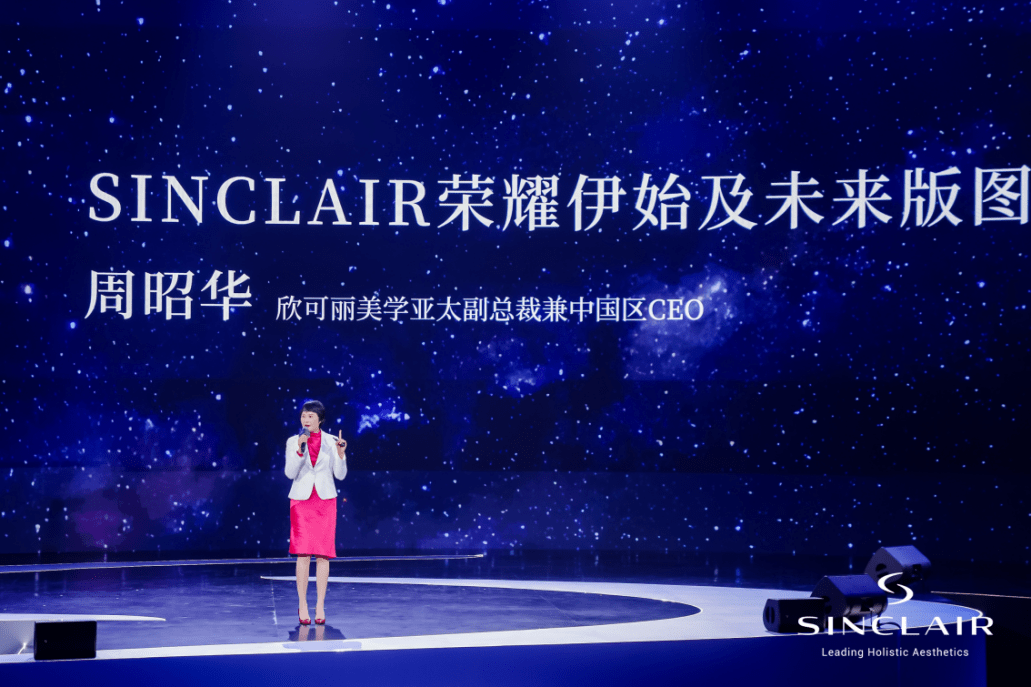 A woman standing on a stage with a large digital screen behind her displaying the name 'SINCLAIR' and Chinese characters, with a starry night background. There are speakers on the stage and she is dressed in a pink skirt, white blazer, and red blouse.