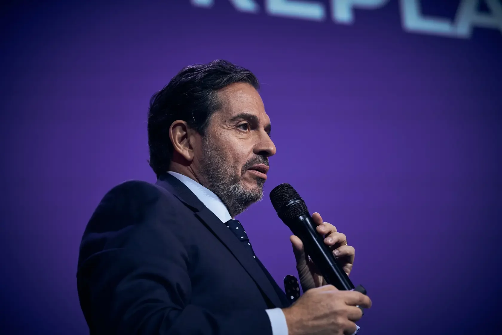 A middle-aged man with dark hair and a beard, dressed in a dark suit and tie, speaking into a black handheld microphone against a purple background.
