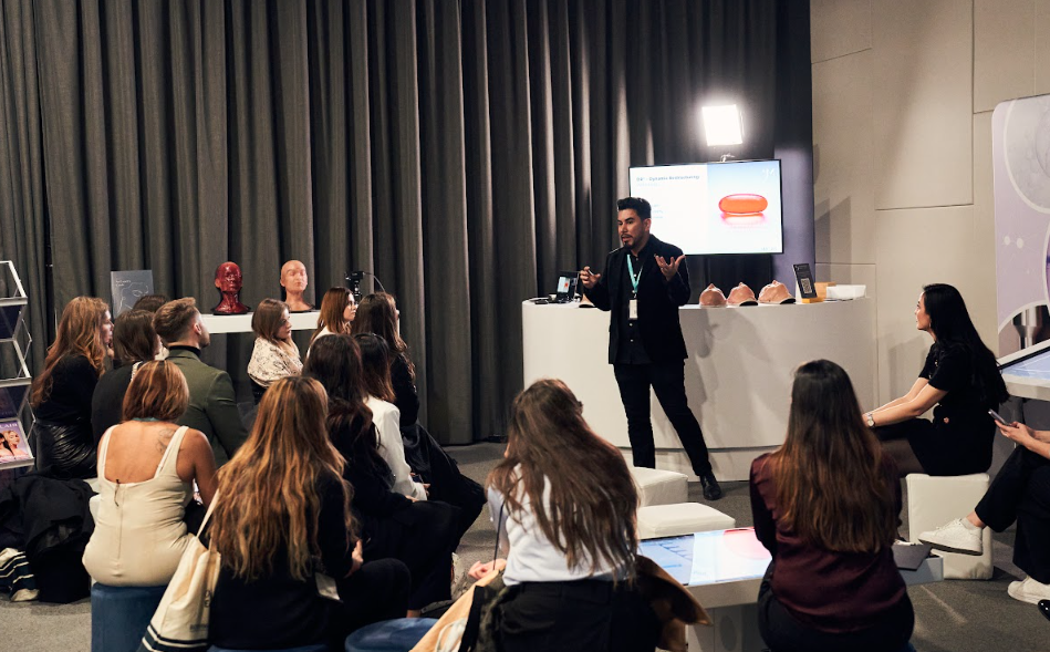 A man giving a presentation to an audience in a conference room with a screen, props, and a white counter in the background.