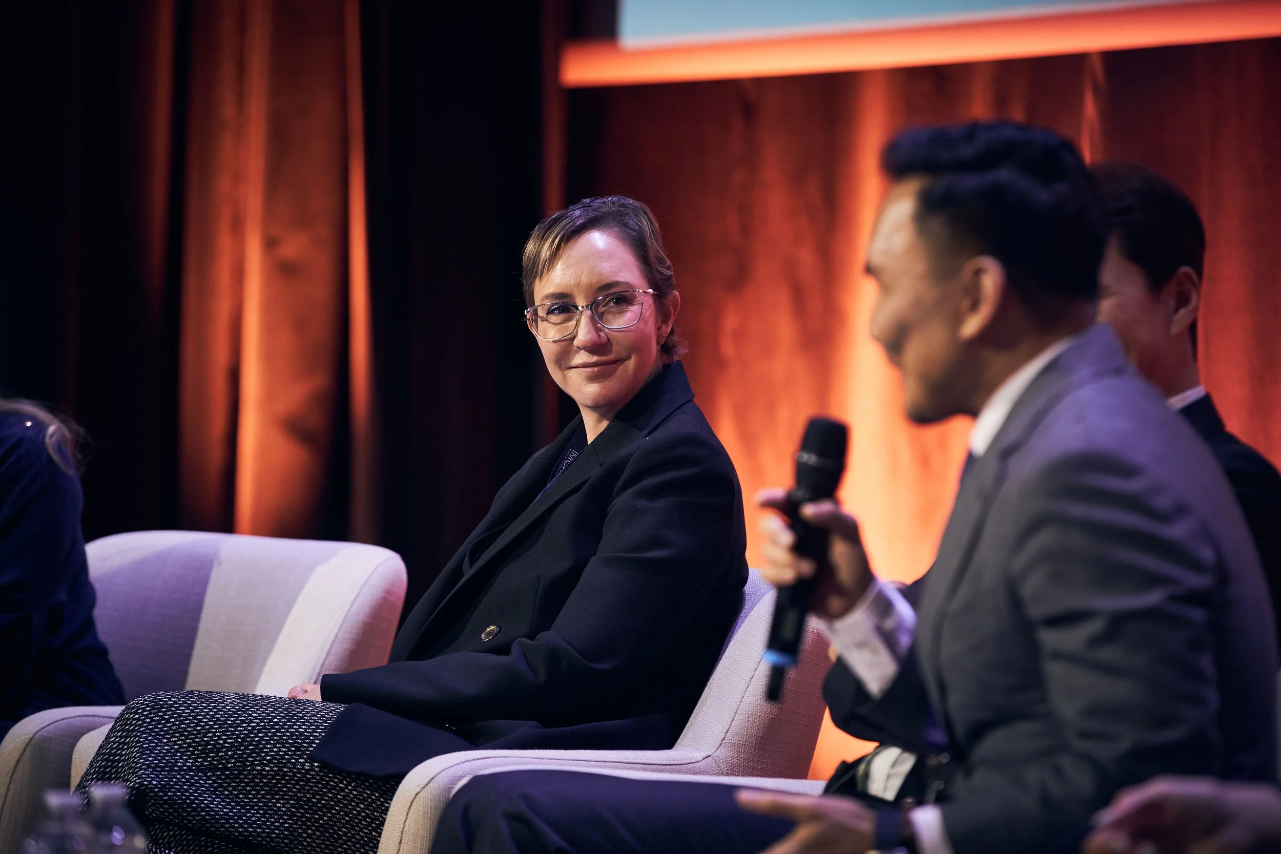 A woman with short hair and glasses sitting on a panel with other people, holding a microphone in a discussion setting with a backdrop of wood paneling and dim lighting.