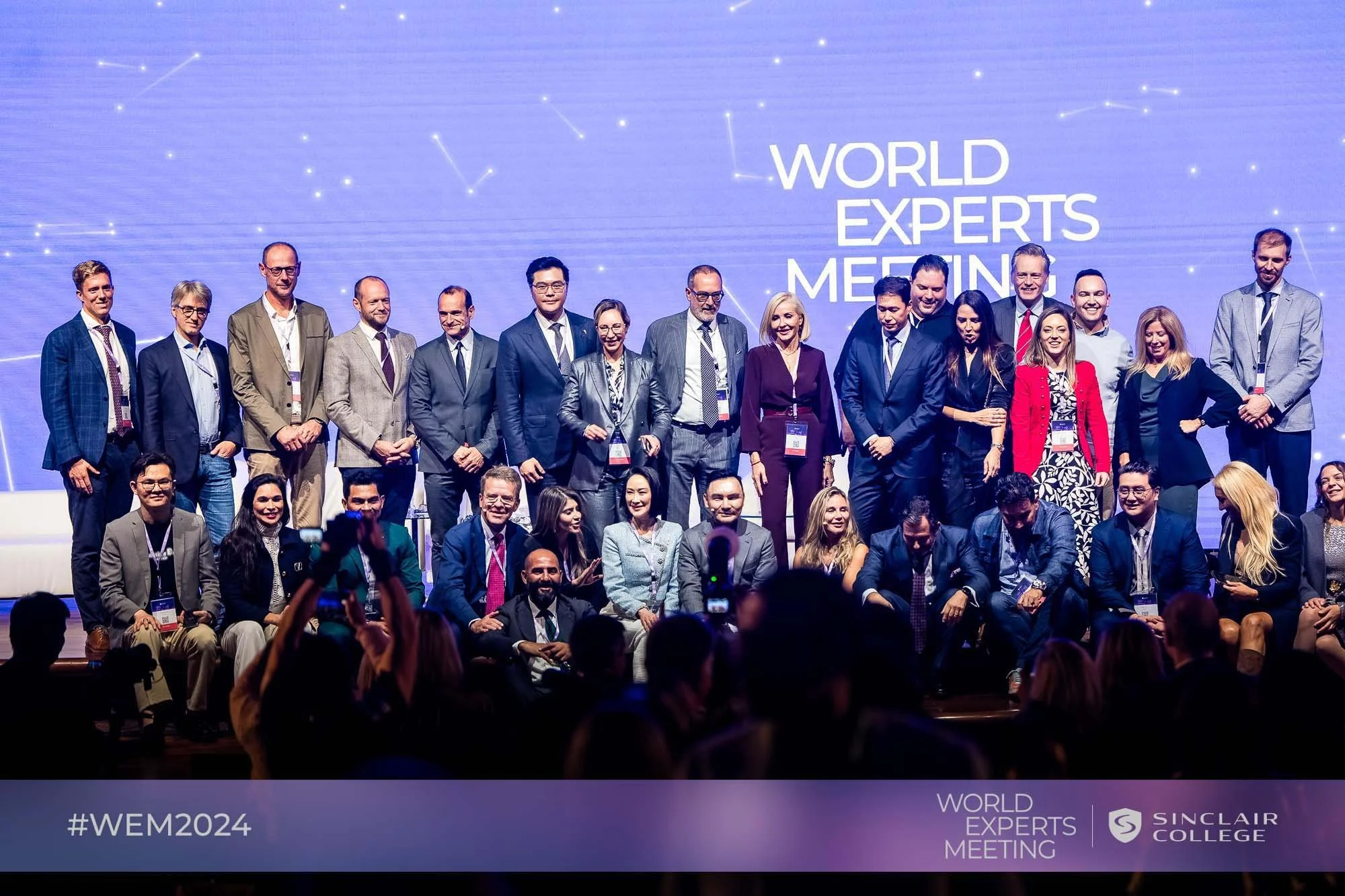 A group of diverse professionals, both men and women, standing on a stage during a conference called 'World Experts Meeting' at Sinclair College. The background displays a blue digital design with the event name and logo, and the bottom left corner has the hashtag #WEM2024.