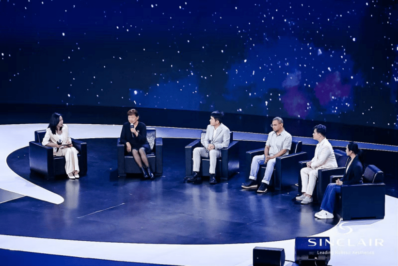 A panel discussion with six people seated on a circular stage against a starry night sky backdrop, with a moderator on the left and five panelists on the right, all engaged in conversation.