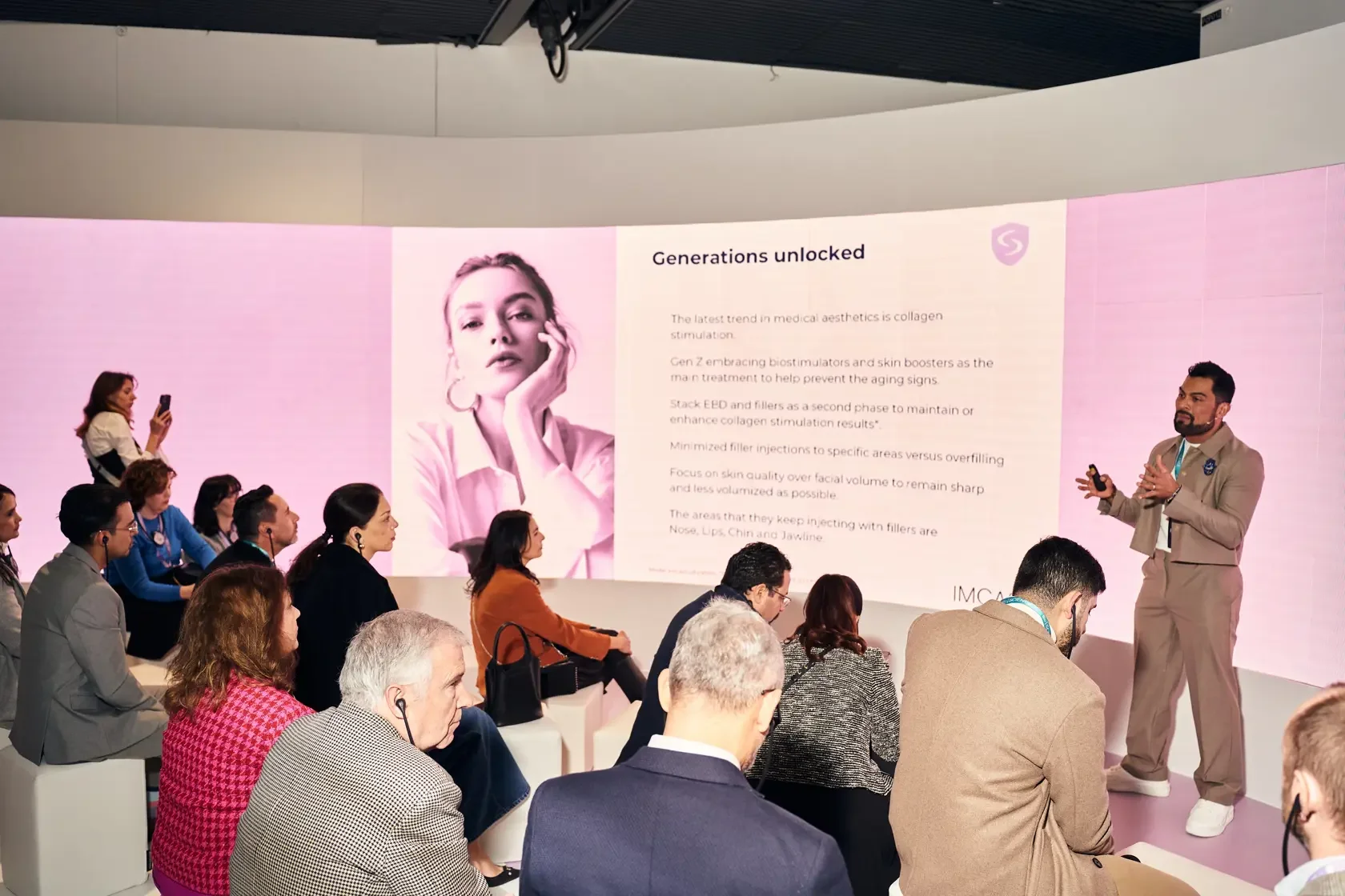 A presenter in beige suit giving a talk on medical aesthetics to an audience. A large screen behind him displays information about generations unlocked in collagen stimulation. The audience is seated, attentively listening, while some take notes or photos.