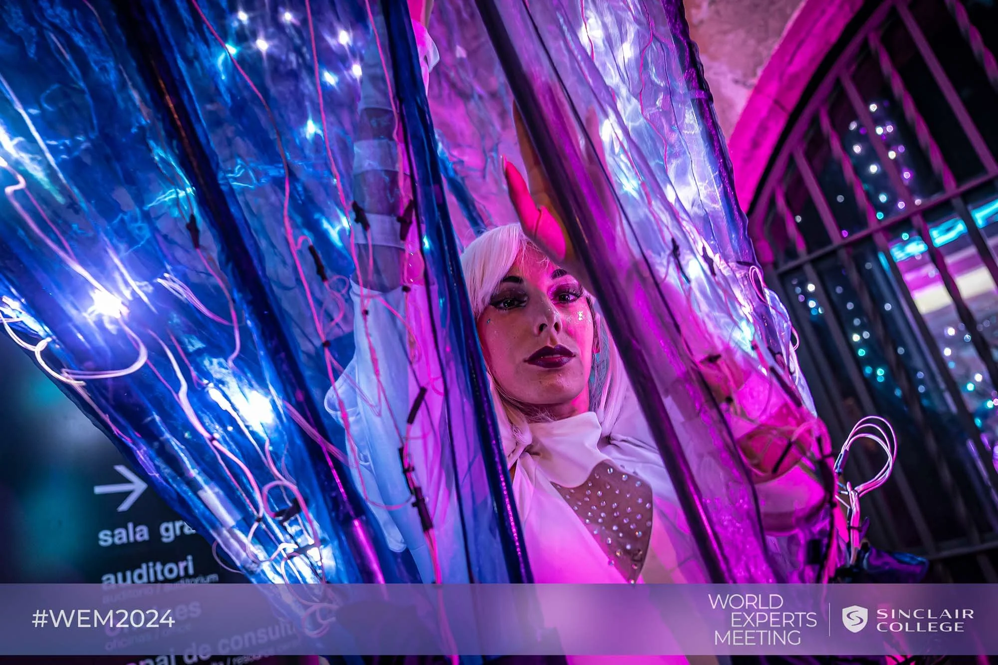 A person with platinum blonde hair and makeup is behind purple and blue LED-lit transparent inflatable tubes at an event. The background shows a dark setting with purple and pink lighting and a railing. The bottom of the image has a banner with text reading '#WEM2024', 'World Experts Meeting', and 'Sinclair College'.