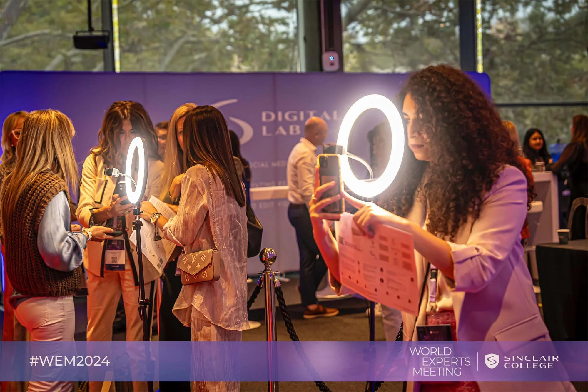 Women gather around a ring light at a professional conference or event, with some holding smartphones. The backdrop features the Sinclair College logo and text indicating a World Experts Meeting and hashtag #WEM2024.