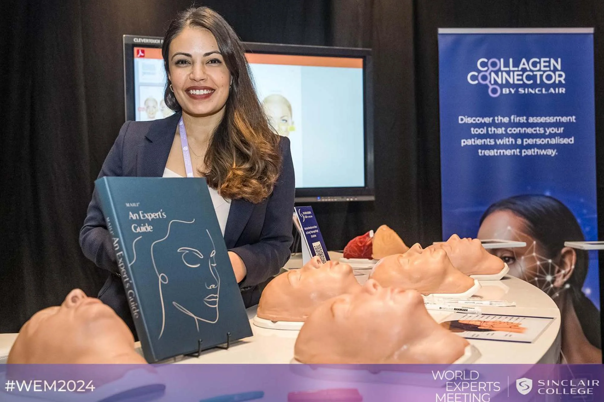 A woman with dark brown hair, wearing a gray blazer, stands smiling at a booth during a conference. She holds a blue booklet titled 'An Expert's Guide' with an abstract face design. There are anatomical models of human heads and brains on the table in front of her. A blue banner in the background reads 'Collagen Connector by Sinclair' and explains the product's purpose. The event is the World Experts Meeting 2024.