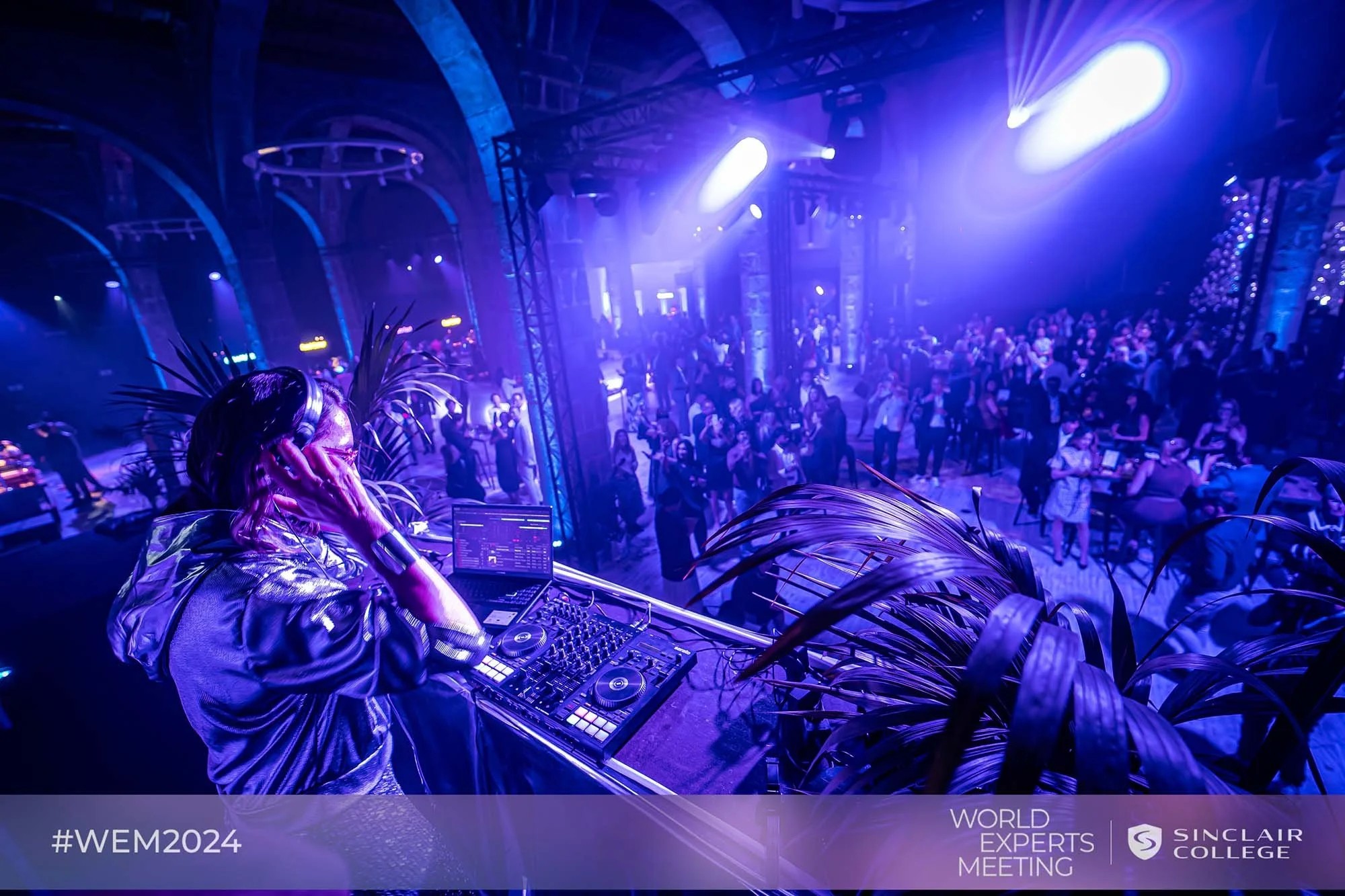 A DJ wearing headphones and a metallic jacket is performing in a nightclub with a large crowd of people dancing, illuminated by vibrant purple and blue lighting, at the World Experts Meeting event sponsored by Sinclair College in 2024.