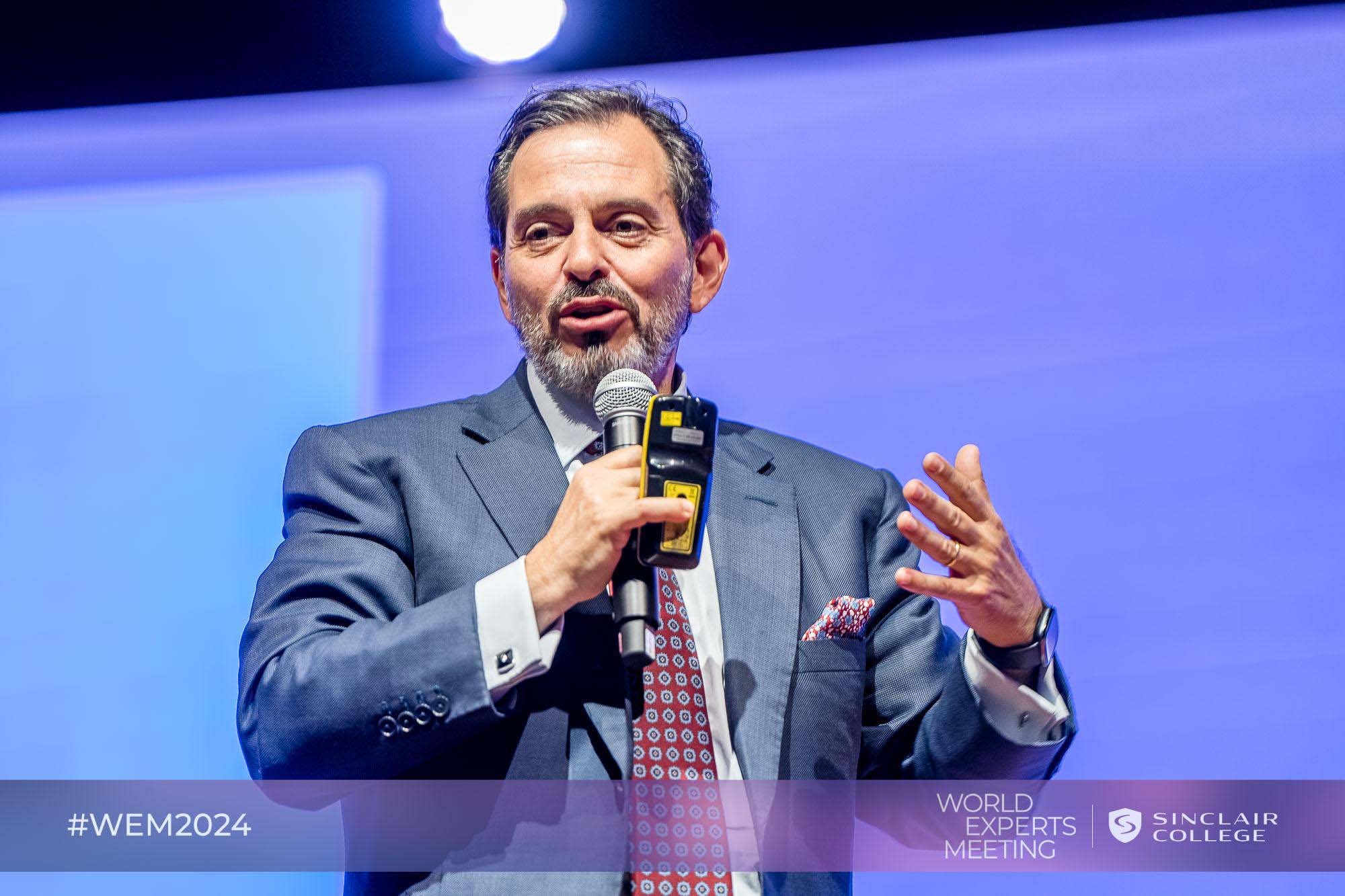 A man in a suit and tie speaking into a microphone during an event with a blue background. There is a banner at the bottom with #WEM2024, WORLD EXPERTS MEETING, and Sinclair College logo.