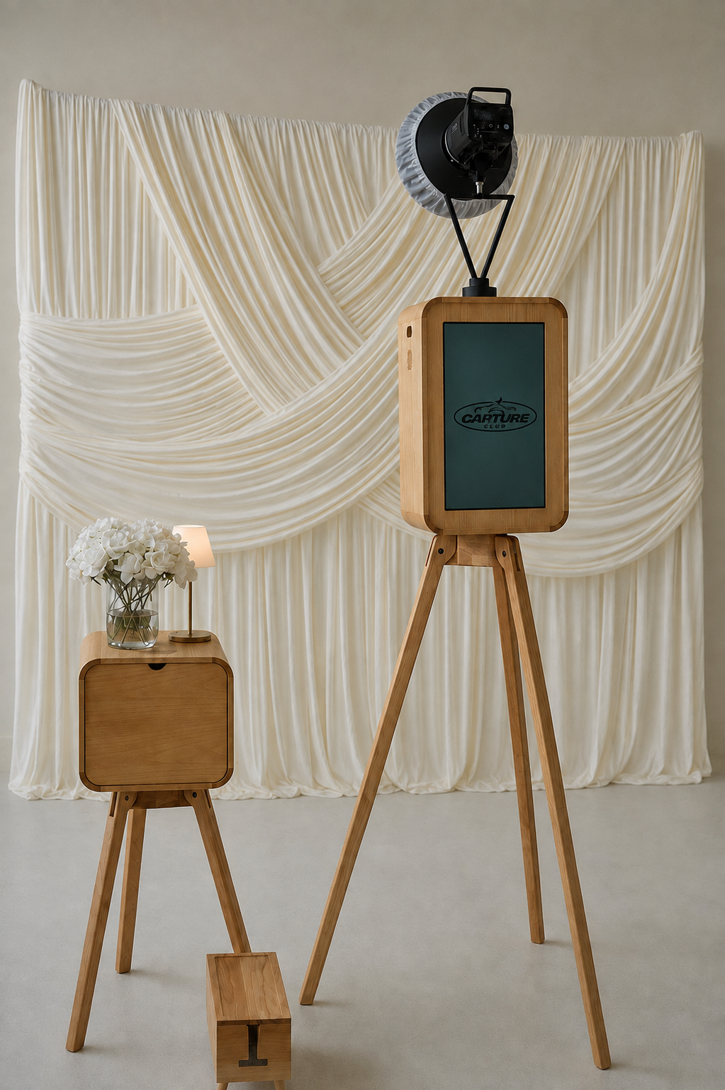 A photo booth setup with a wooden tripod photo booth, a backdrop of cream-colored draped fabric, a small wooden side table with a vase of white flowers and a small lamp, and a large reflector with studio lighting at the top.