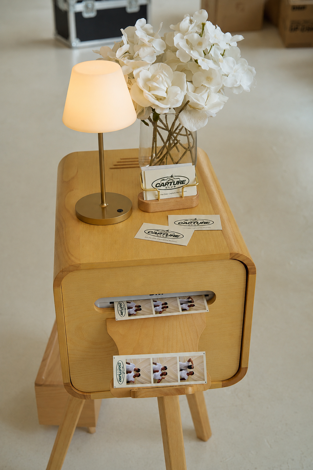 A wooden table with a small lamp, a glass vase filled with white flowers, and promotional cards and stickers for Capturing Club, with some printed photos hanging below.