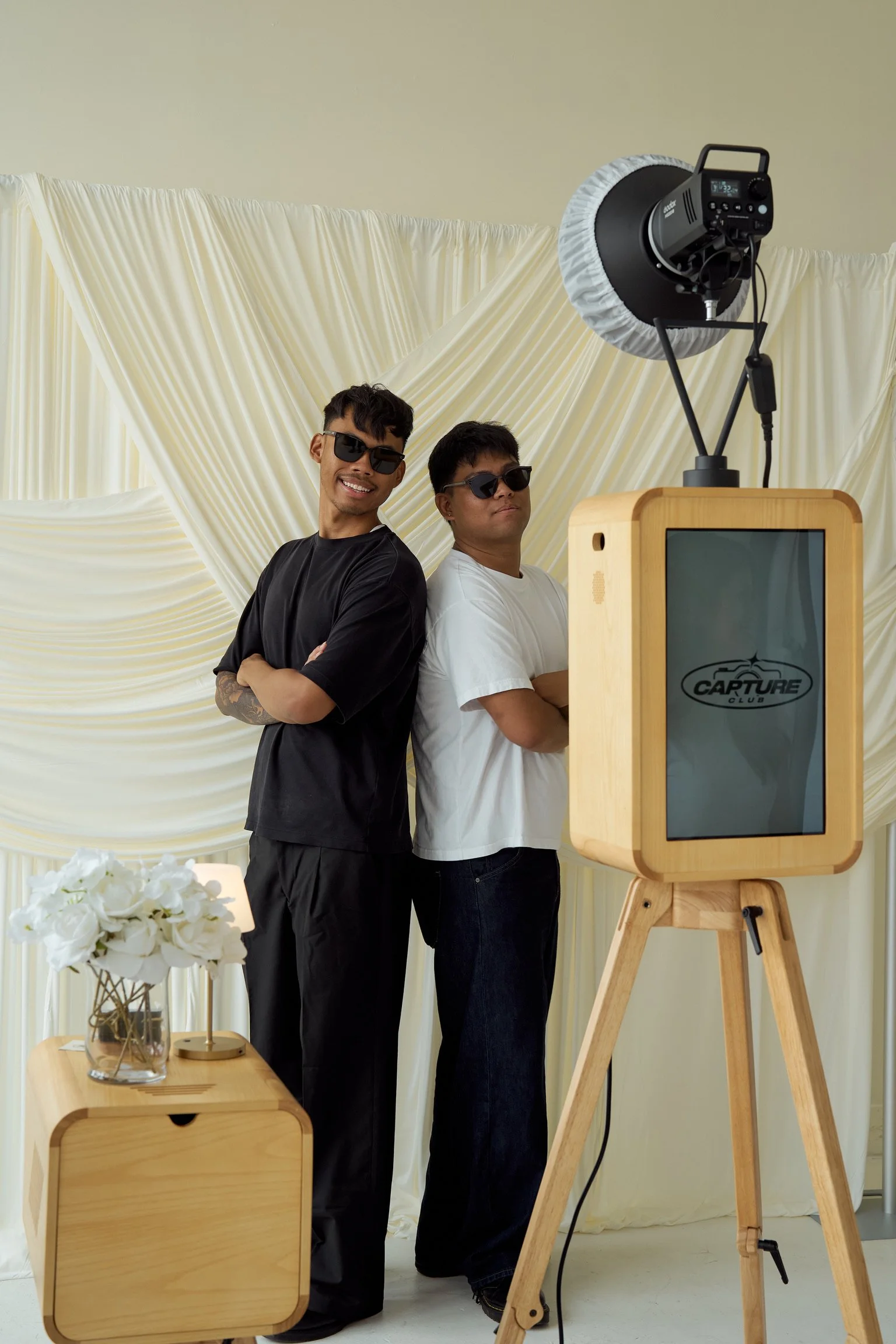 Two men wearing sunglasses stand back to back in front of a neutral backdrop, with a photo booth setup and a bouquet of white roses on a small wooden table in the foreground.