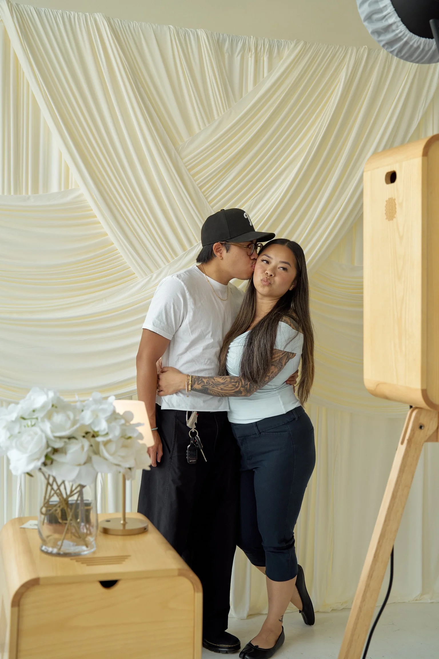 A couple takes a selfie in front of cream-colored draped fabric, with the man kissing the woman's cheek and both posing with puckered lips. The woman has tattoos on her arm and is wearing black shoes, while the man wears glasses, a black baseball cap