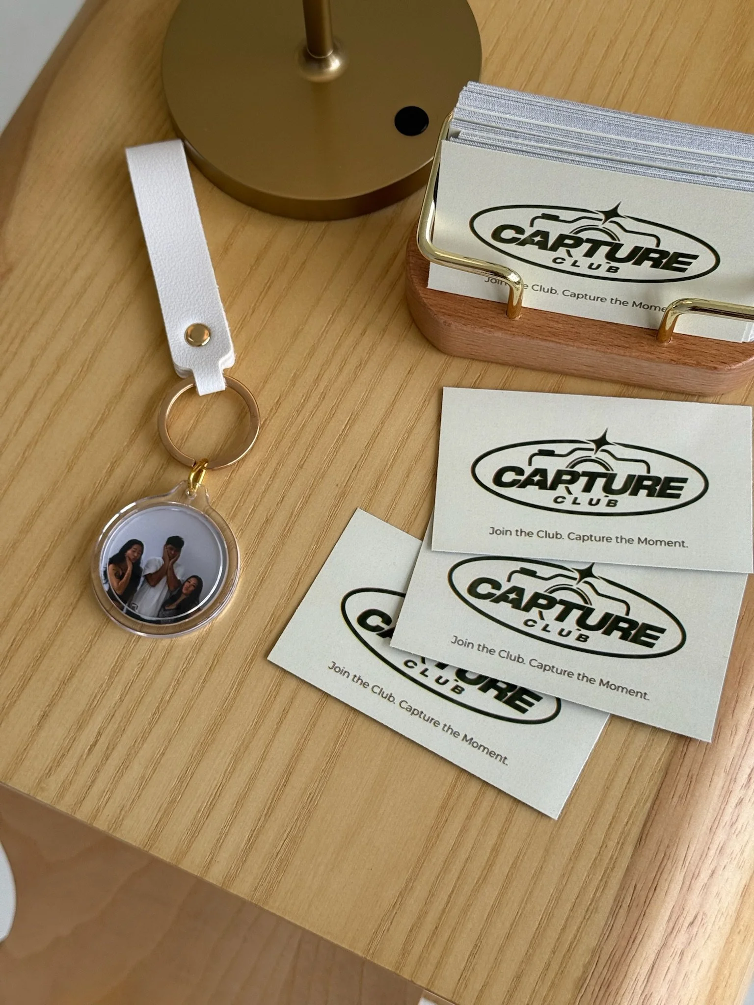 Business cards and a keychain with a photo of three people on a wooden table near a gold-colored lamp base.