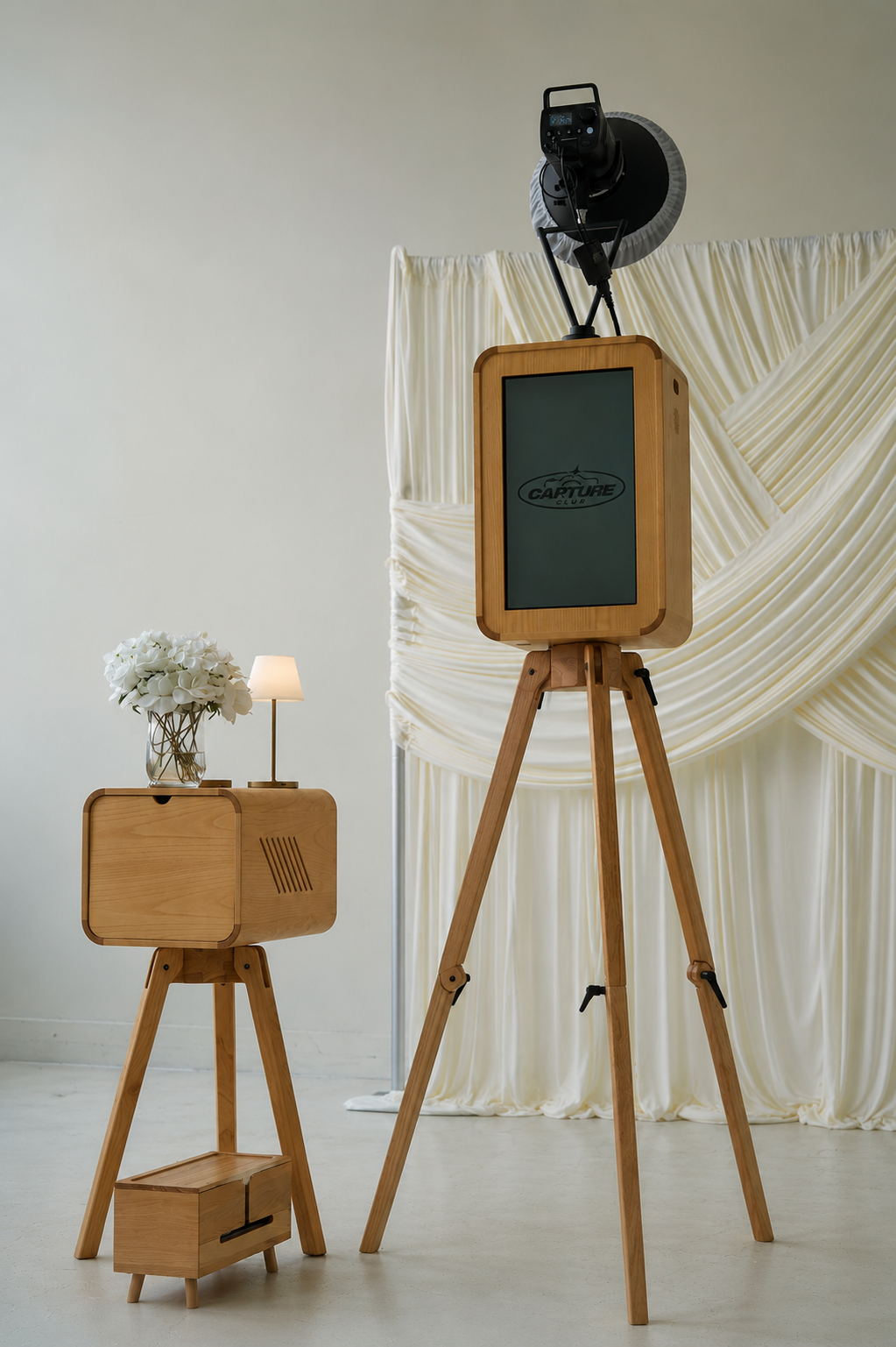 Photograph of a vintage film camera on a tripod, a wooden picture with a screen displaying the logo "Capture Club," a small wooden cabinet with a lamp and a vase with white flowers, and a cream-colored fabric backdrop.
