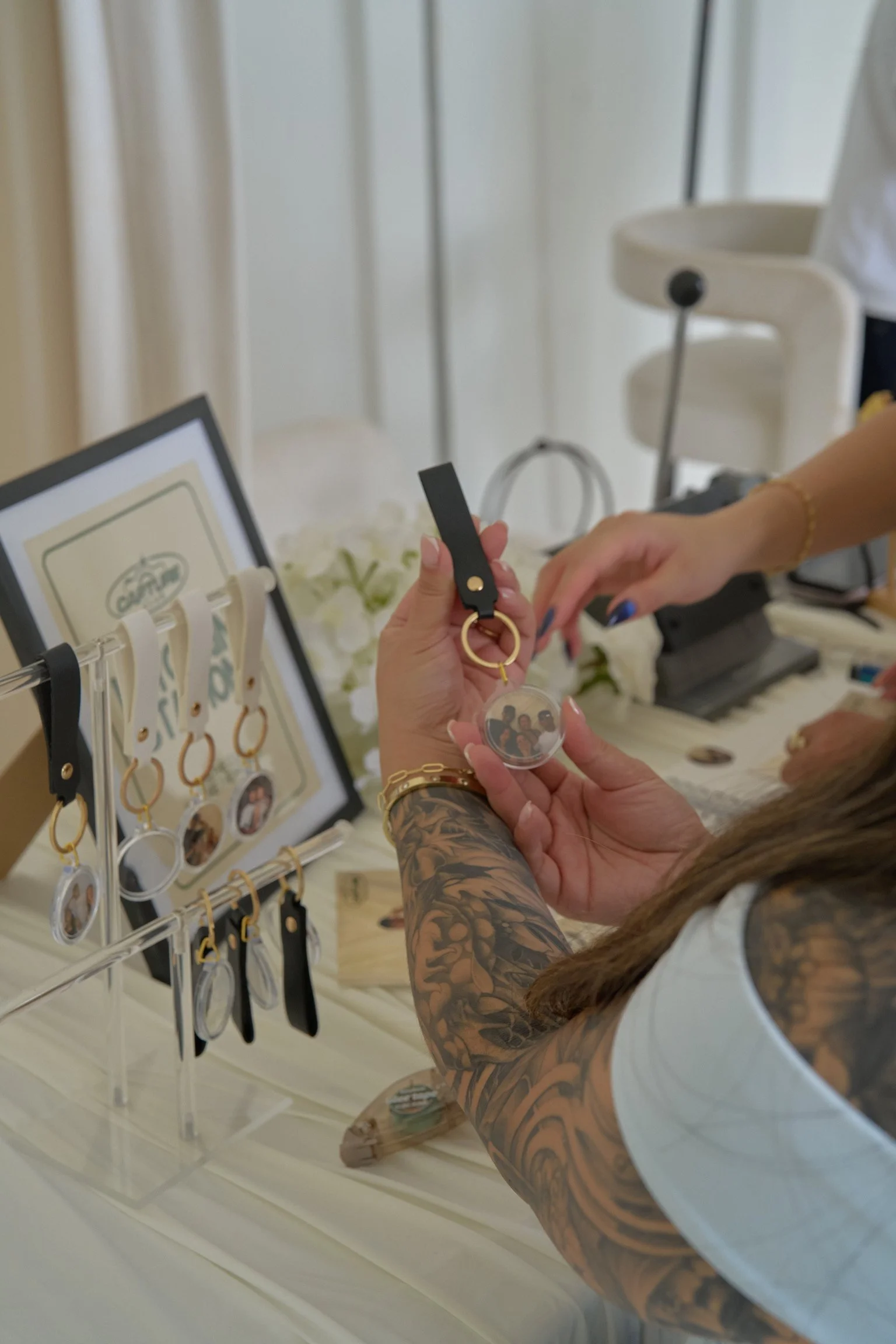 Close-up of a woman with detailed tattoos on her arm, handing a keychain with a photo to another person. In the background, a display stand with various keychains and picture frames, and a blurred white high chair. The scene appears to be at a booth 