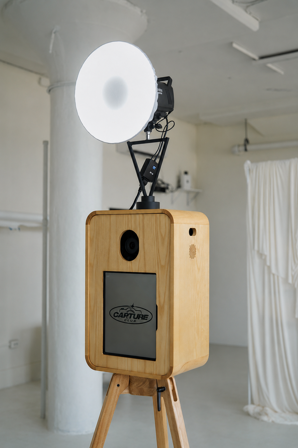 A photo of a wooden photo booth with a large camera lens on a tripod, a digital display that says 'CAPTURE,' and a light source mounted above it, inside a light-colored room with a white curtain on the right side.