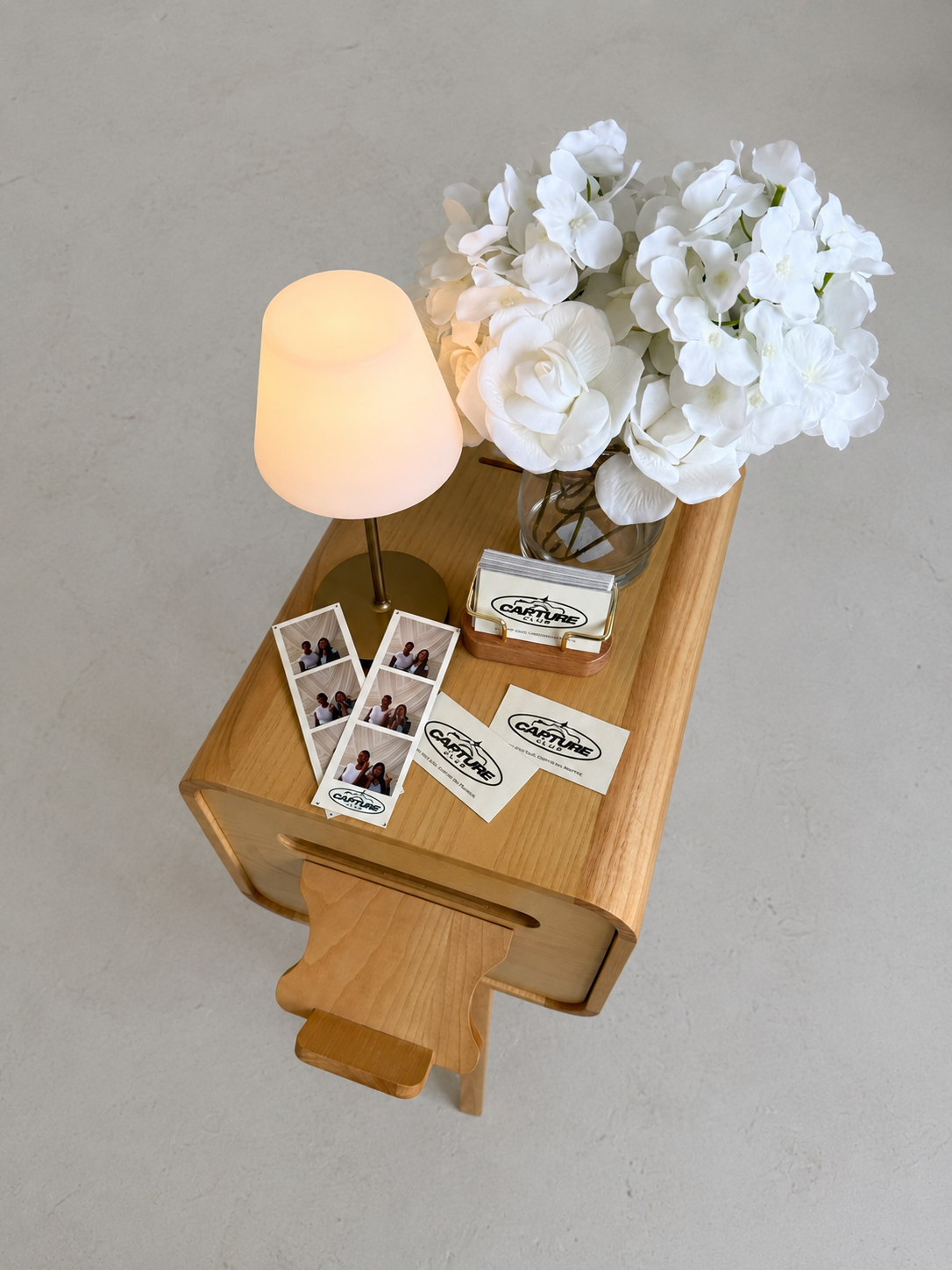 Wooden table with a white flower bouquet in a glass vase, a small lamp, several contact sheets with photographs, and business cards for Capture Club.