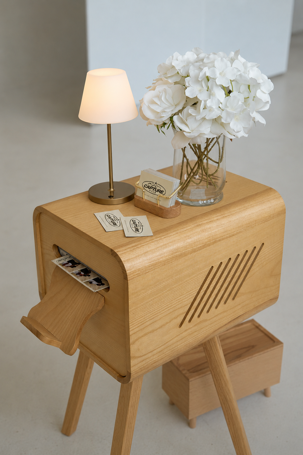 A wooden table with a small lamp, a glass vase of white flowers, and business cards with a wooden cardholder. There are also two small printed photos or cards on the table.