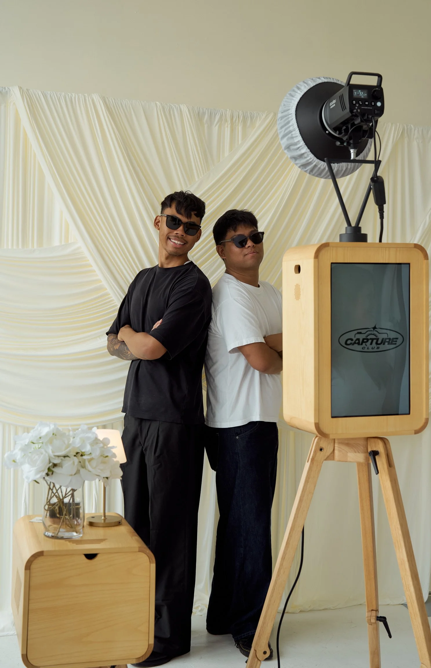 Two men wearing sunglasses standing back-to-back in front of a photo booth, with a wooden photo booth and a backdrop of cream-colored drapes. A table with a bouquet of white flowers is also visible.