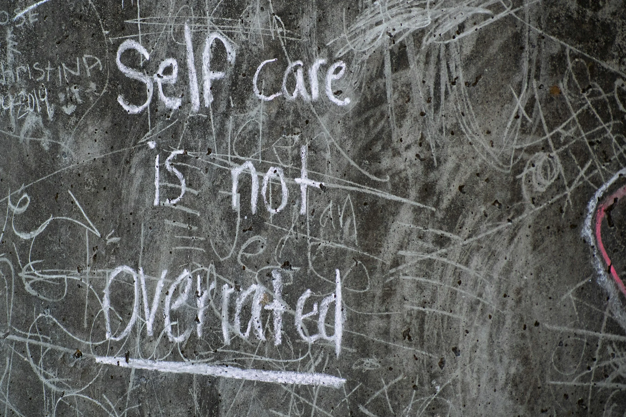 A concrete wall covered in white chalk graffiti and scribbles with the words 'Self Care' written prominently in the center.
