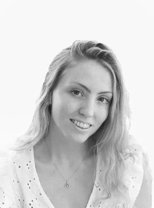 Black and white portrait of a young woman with light hair, smiling, wearing a white blouse with eyelet details and a necklace.