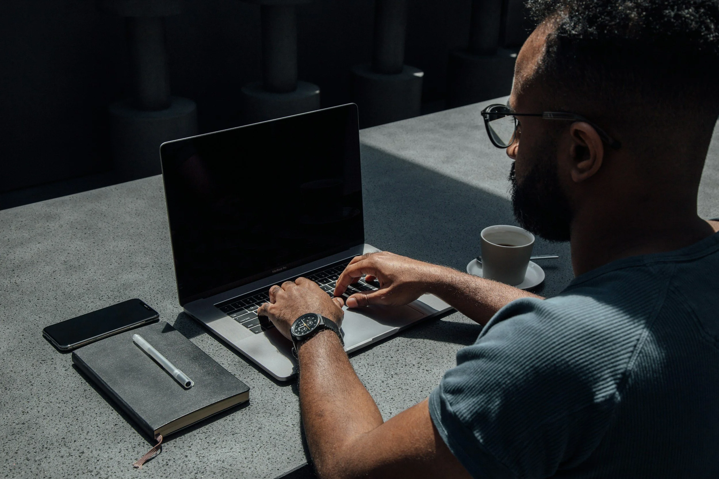 A man with short dark hair and glasses working on a laptop at a desk outdoors, with a cup of coffee, a smartphone, a pen, and a closed notebook nearby.