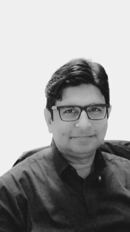 Black and white photo of a man wearing glasses, smiling, and dressed in a dark shirt.