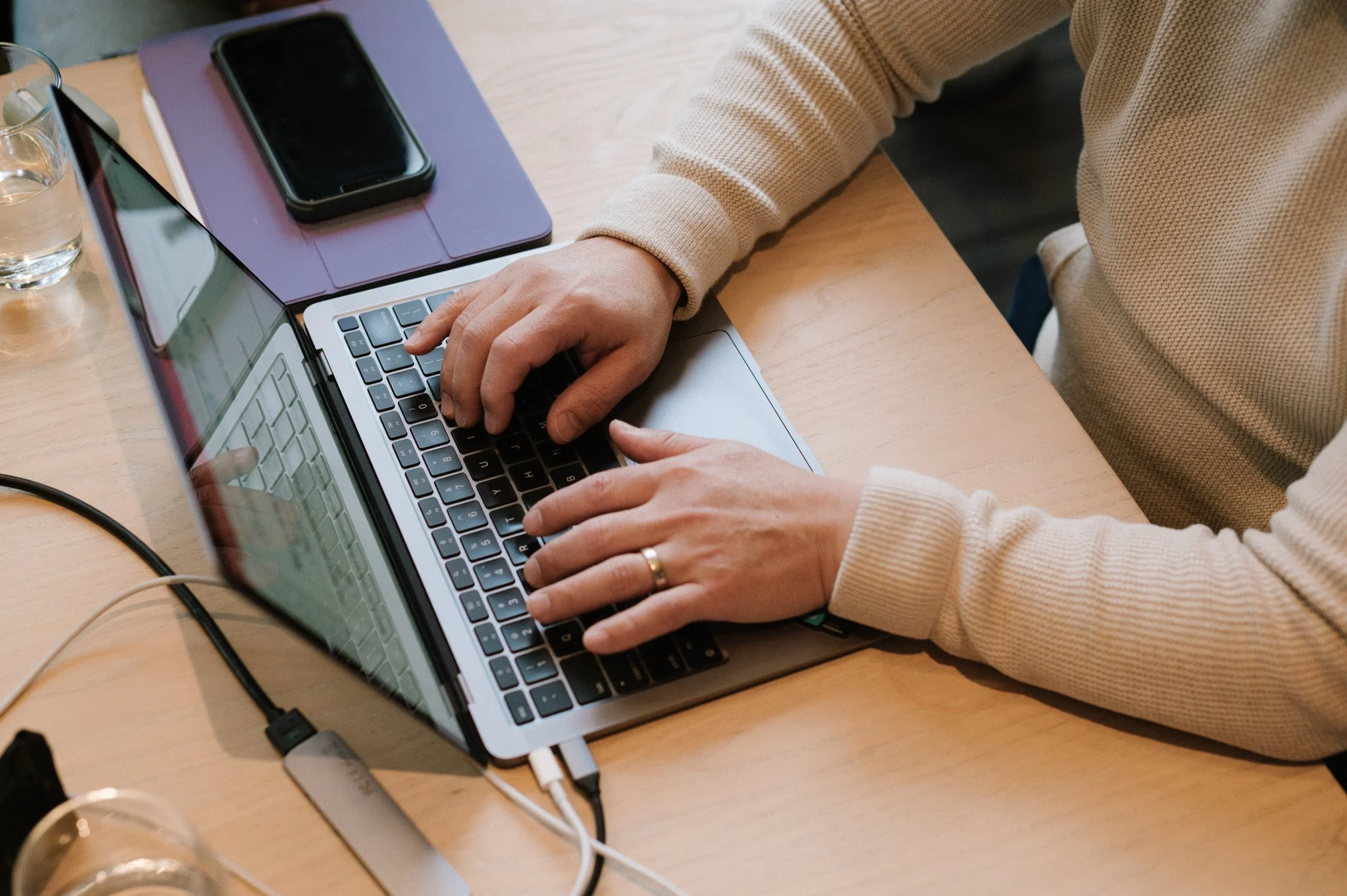 Person using a laptop at a wooden desk with a smartphone, a purple notebook, and glasses of water nearby.