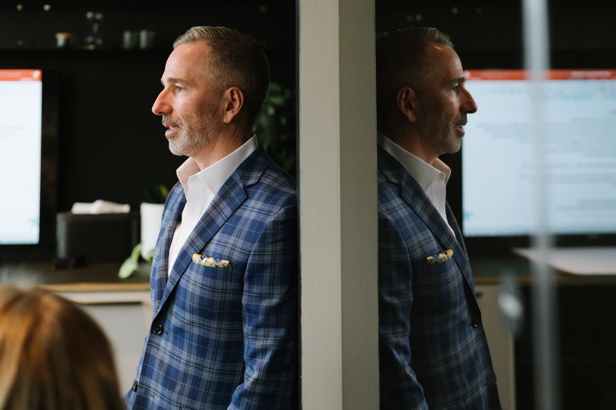 A man with gray hair and a beard wearing a blue checkered blazer and white shirt, standing in profile and leaning against a wall, with his reflection visible in a mirror.