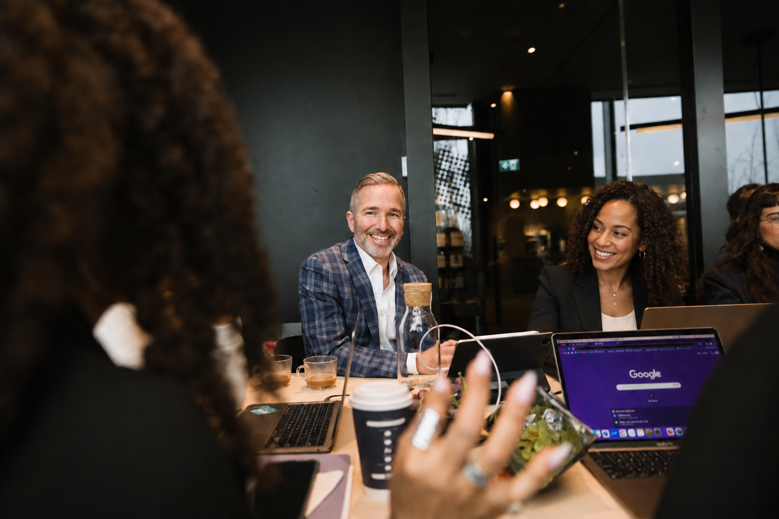 Group of diverse professionals having a meeting at a modern conference room with laptops, coffee cups, and a water bottle, smiling and engaging with each other.