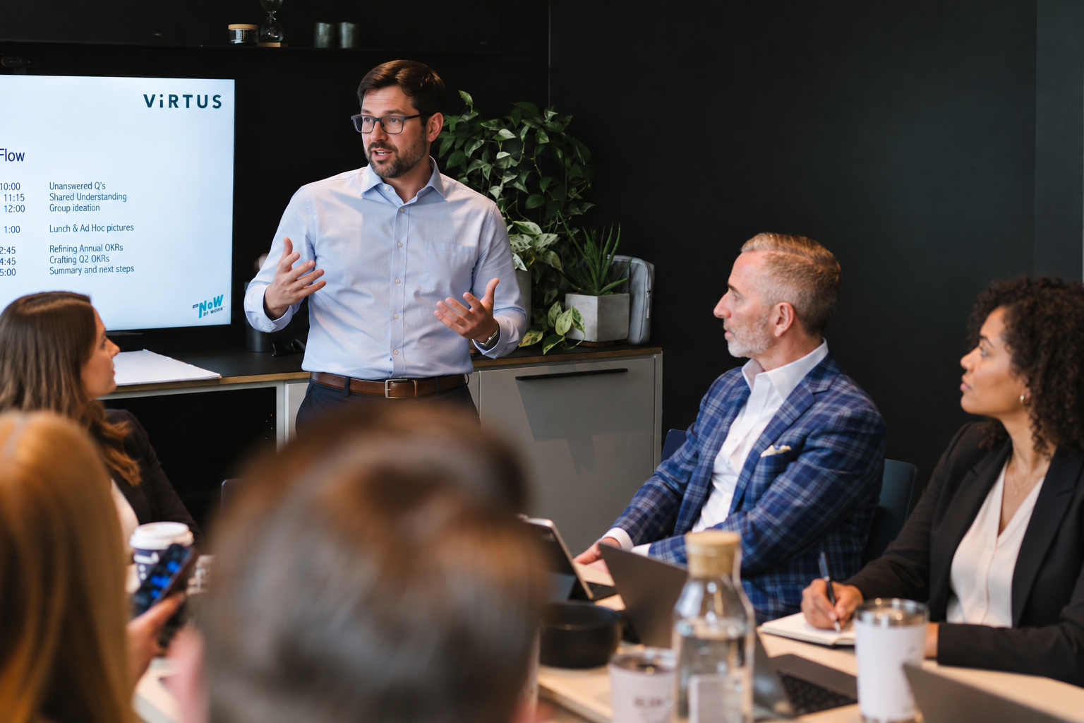 Man giving a presentation to a group in a conference room with a large screen displaying a schedule, several attendees seated and listening, with plants on a cabinet in the background.