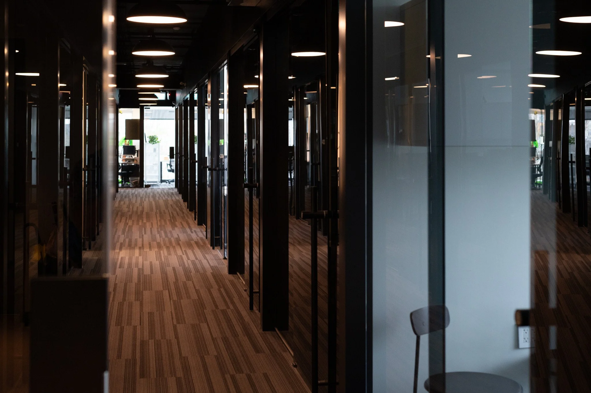 Corridor of an office building with glass doors on either side and ceiling lights. Visible workspaces and windows at the end of the corridor.
