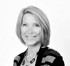 Black and white portrait of a woman with blonde hair, smiling, wearing a patterned top and a necklace.