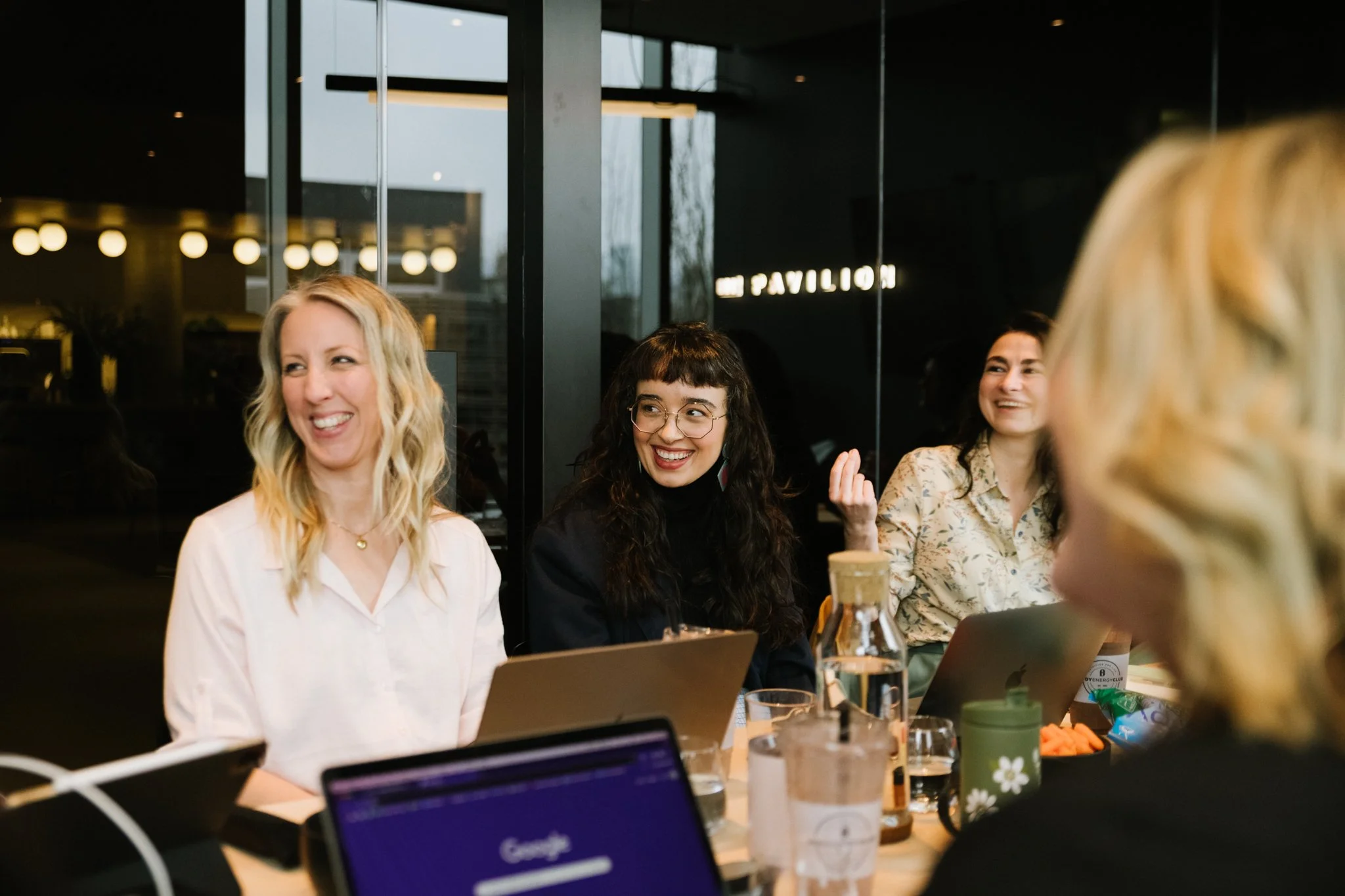 Group of women sitting around a table in a modern, well-lit indoor space, smiling and engaging in conversation.